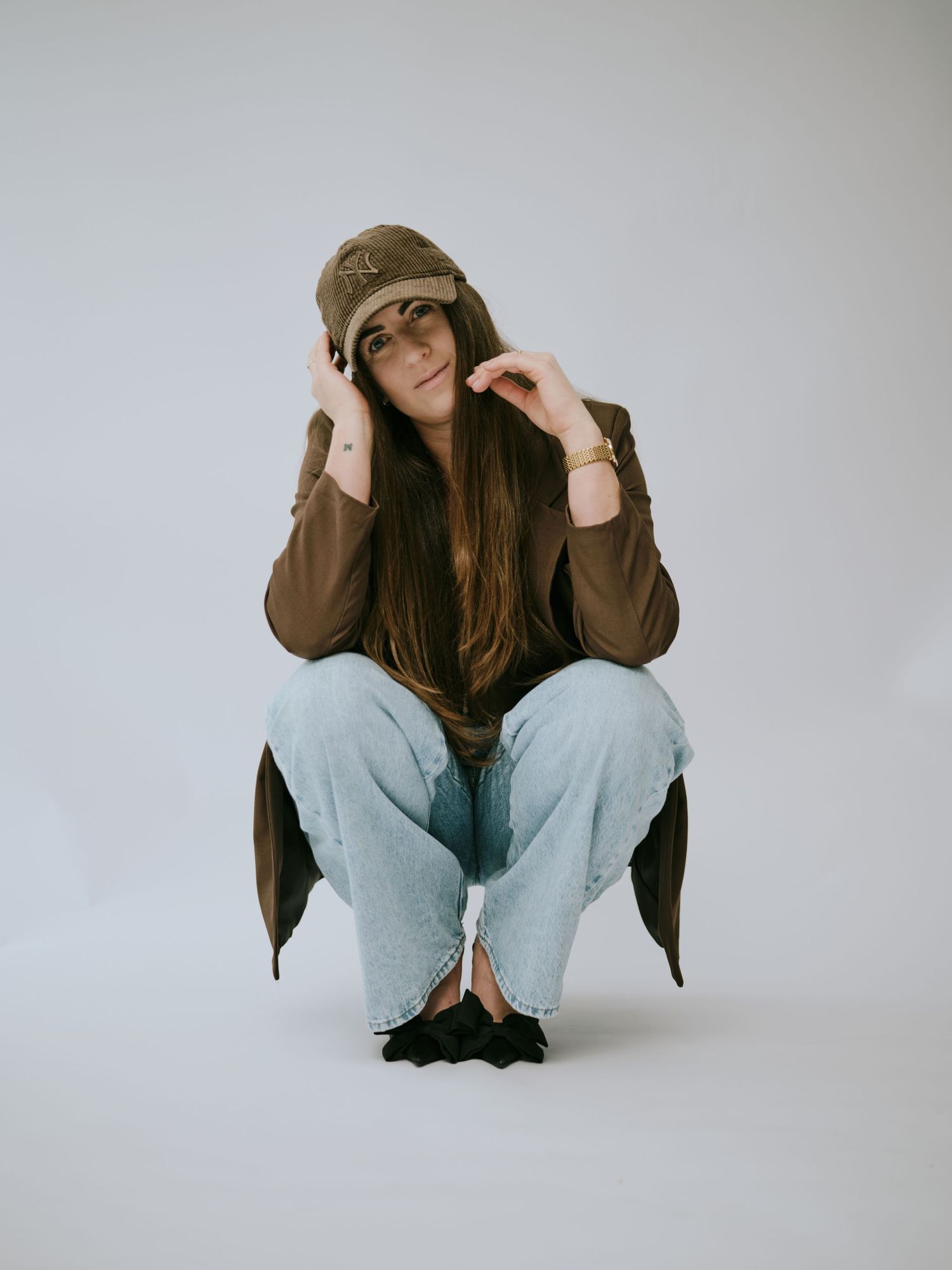 Figure in brown jacket and light jeans crouches in artistic poses against white backdrop in photo series.
