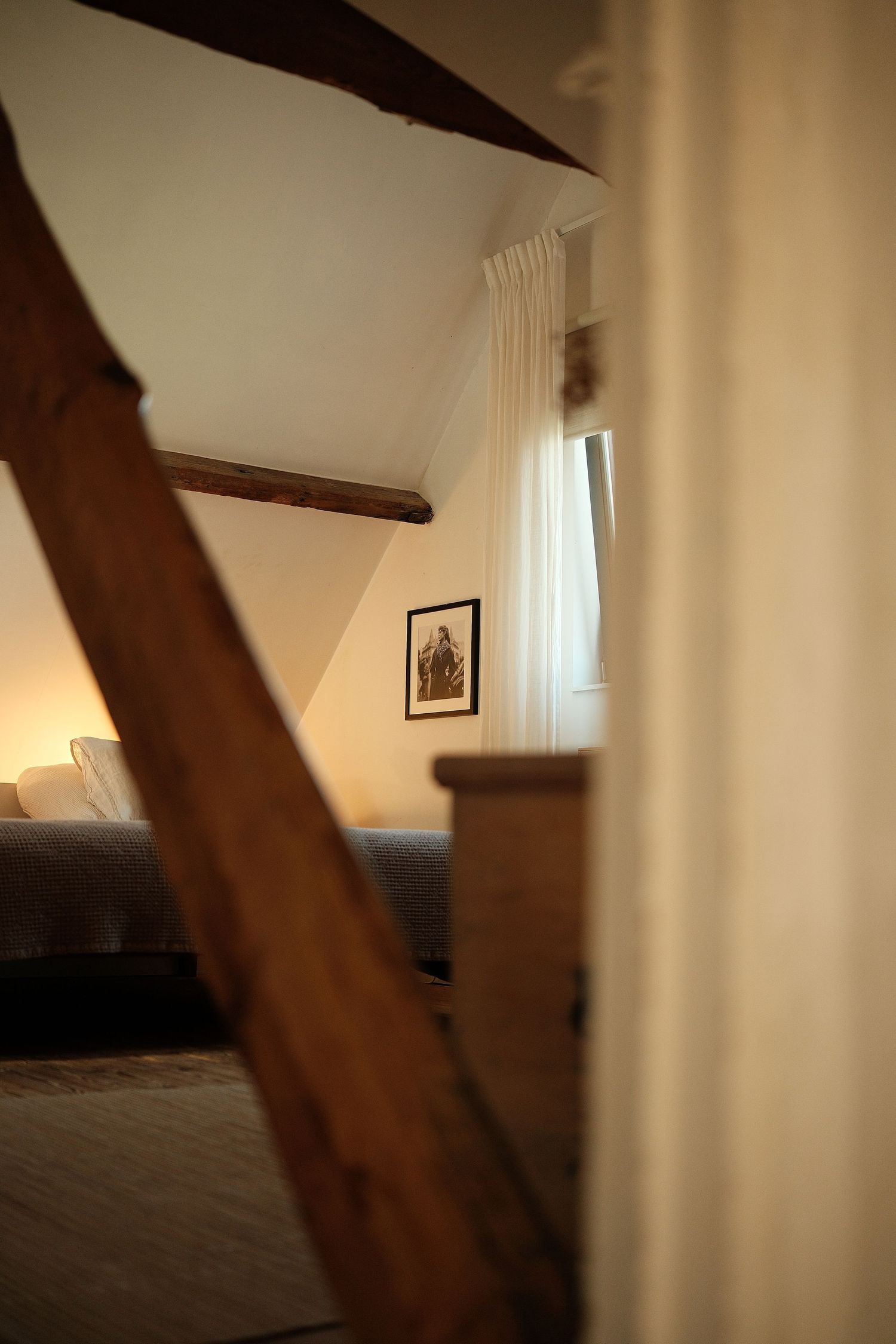 Cozy bedroom interior with exposed wooden beams, white curtains, and natural light streaming through the window.