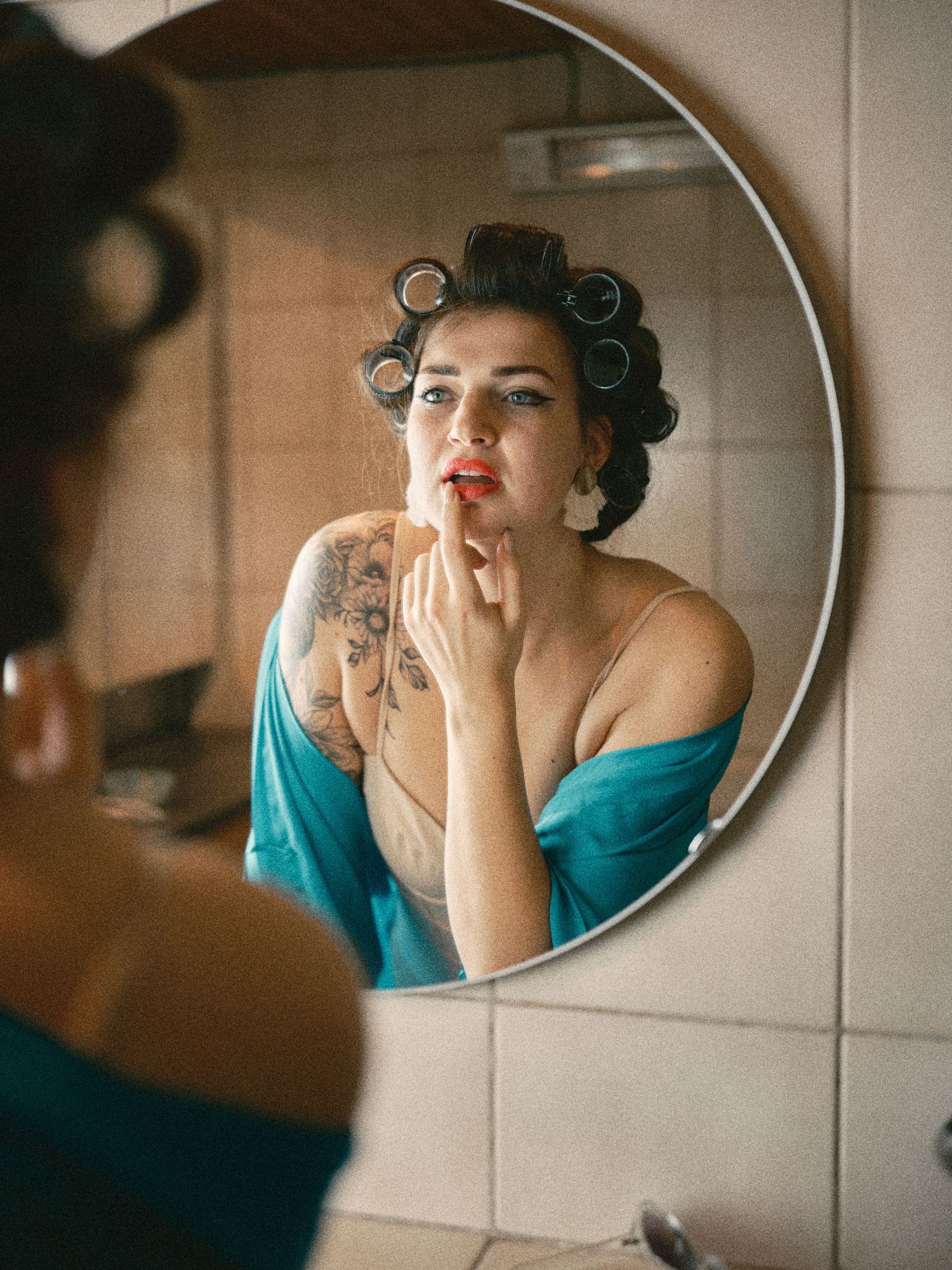 Woman with hair rollers applying red lipstick while looking in a round bathroom mirror, wearing a teal robe.