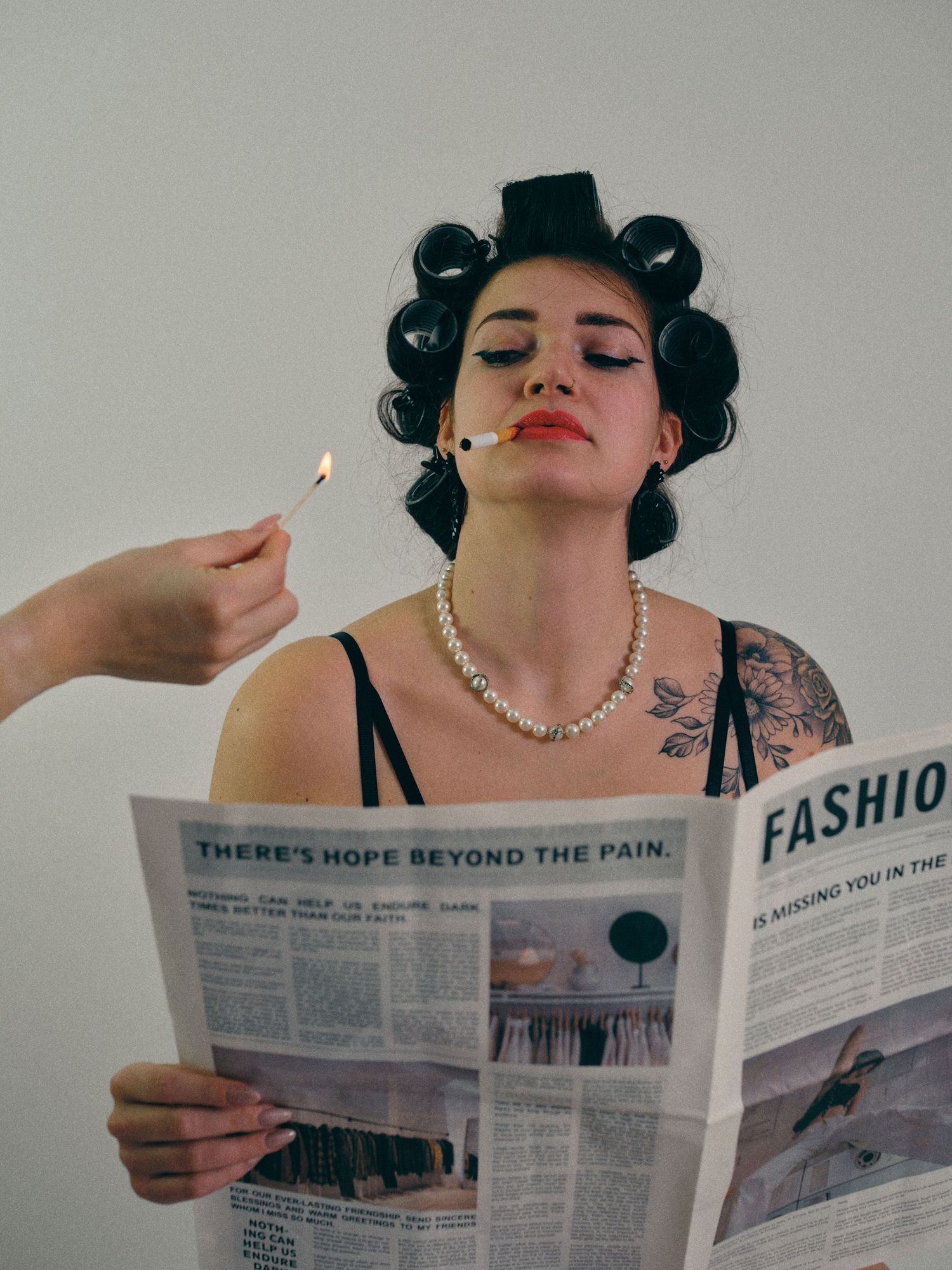 Tattooed woman with hair rollers reads fashion newspaper while someone lights her cigarette, wearing pearl necklace.