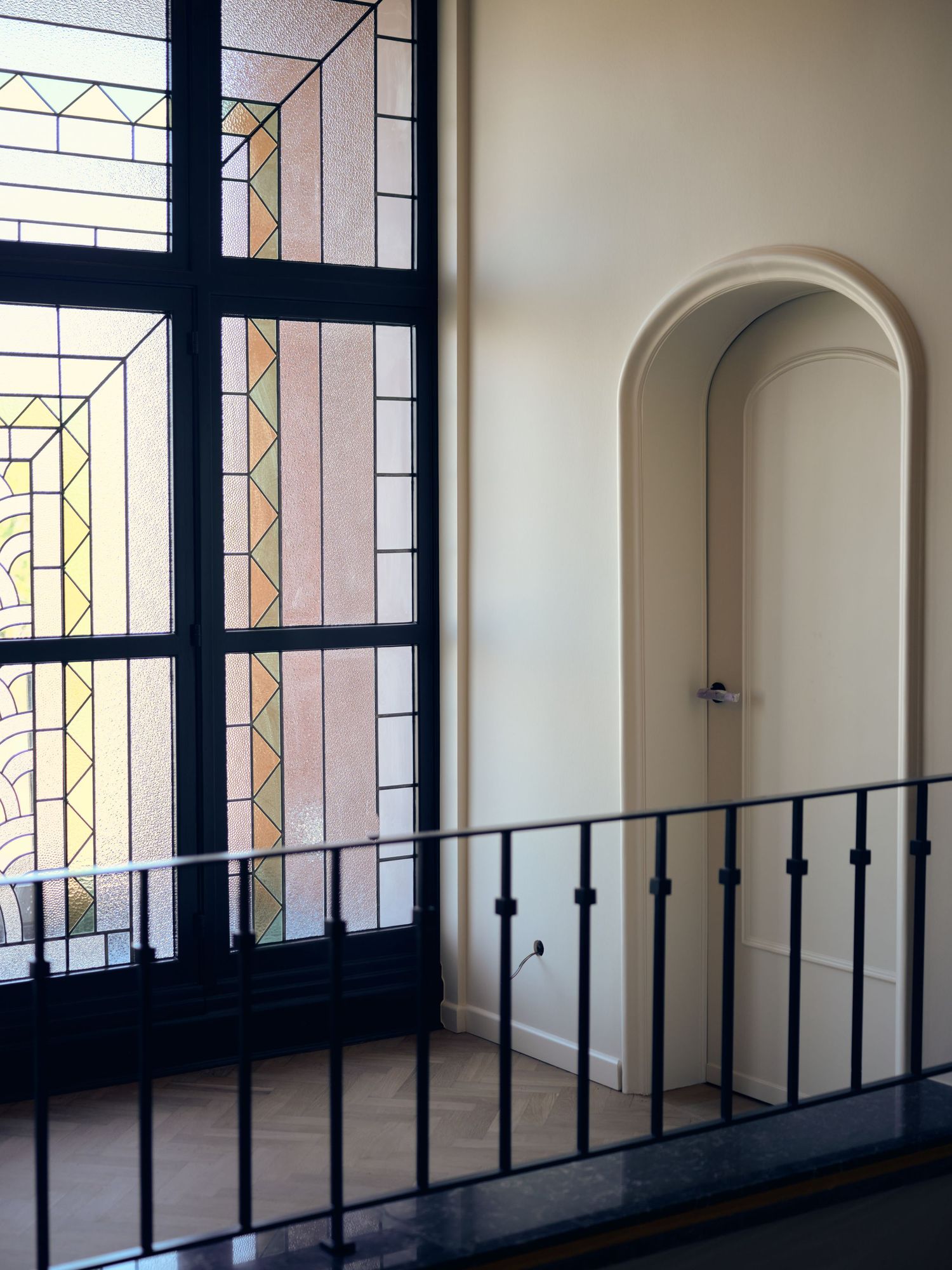 Art Deco stained glass window beside an arched alcove on an elegant interior balcony with wrought iron railing.