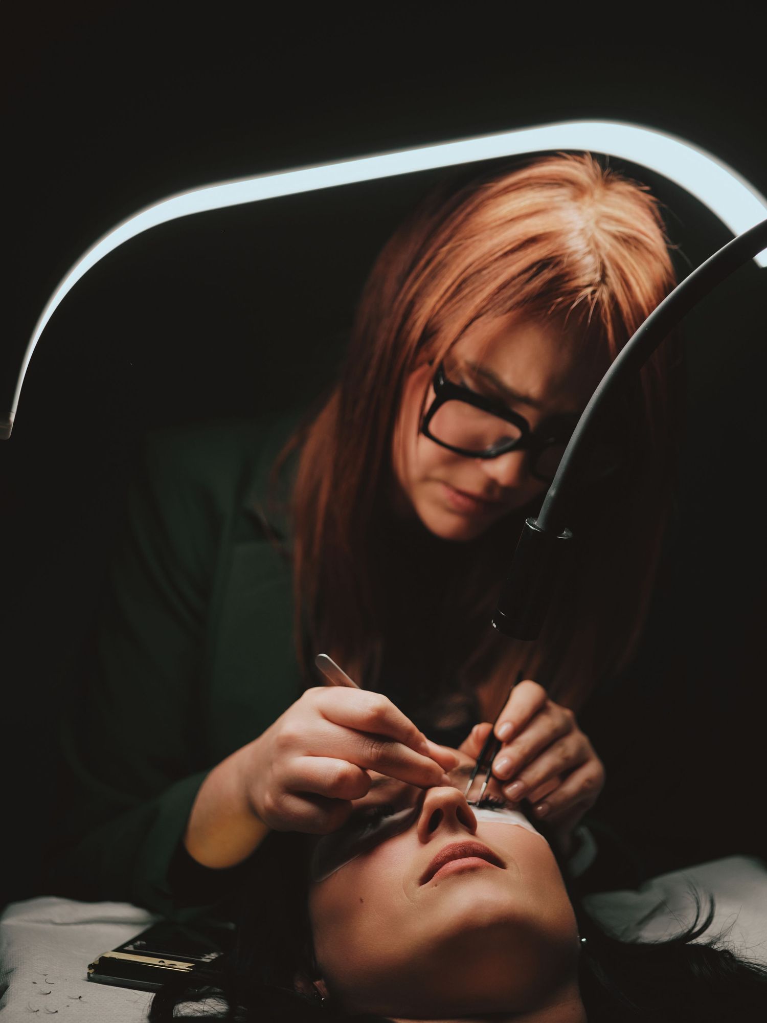 A red-haired esthetician wearing glasses applies eyelash extensions to a client under bright salon lighting.