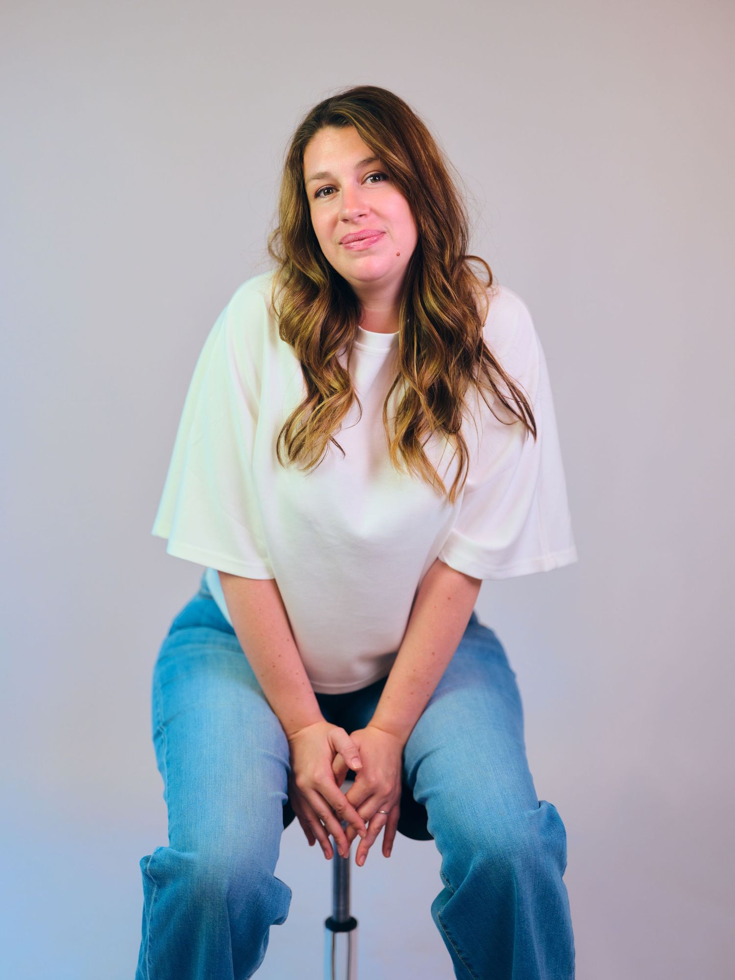 Young woman with wavy brown hair sits on a stool wearing a white top and jeans, smiling at the camera.