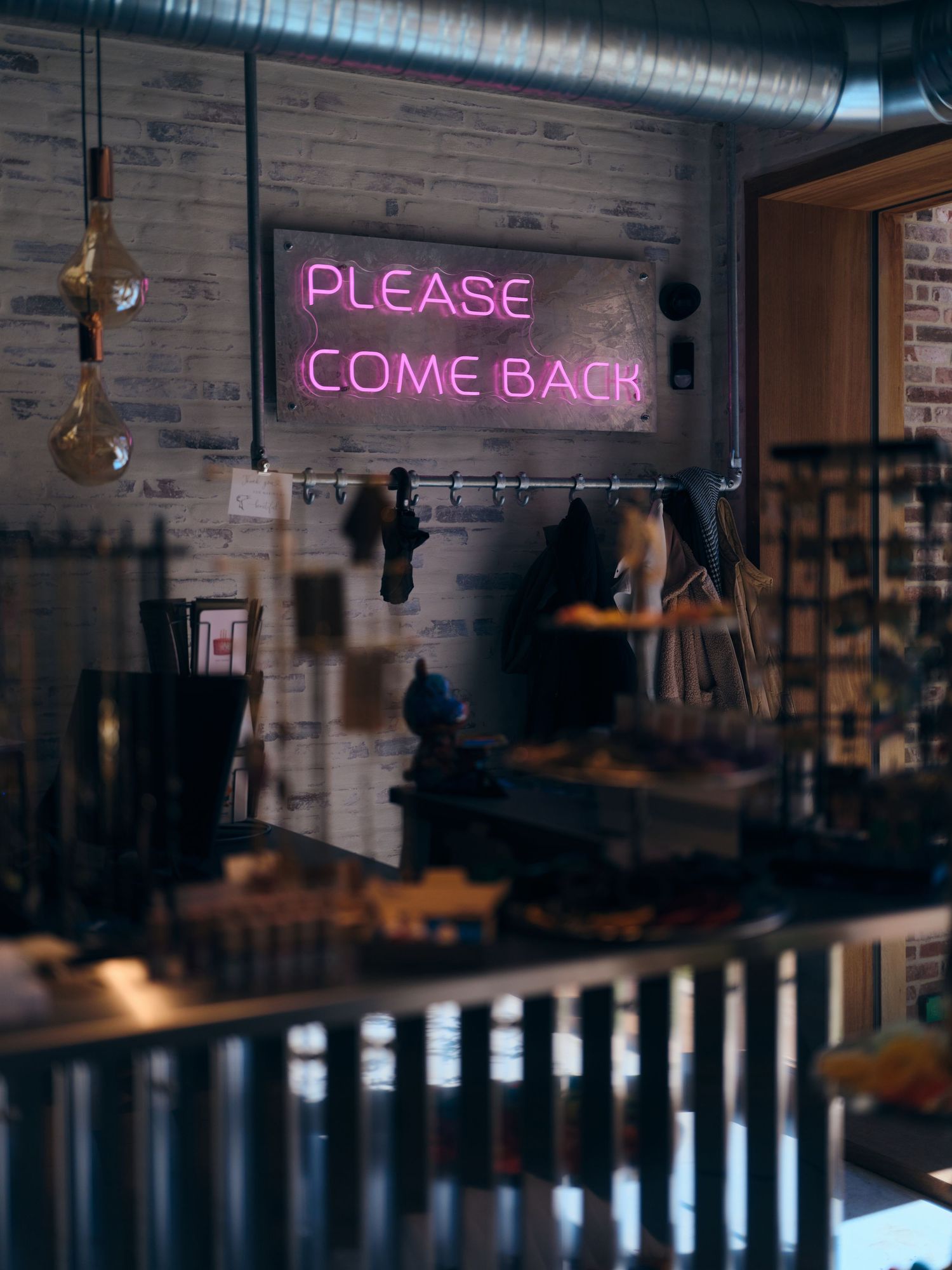 Pink neon sign reading 'Please Come Back' glowing in a dimly lit industrial-style bar or cafe interior.