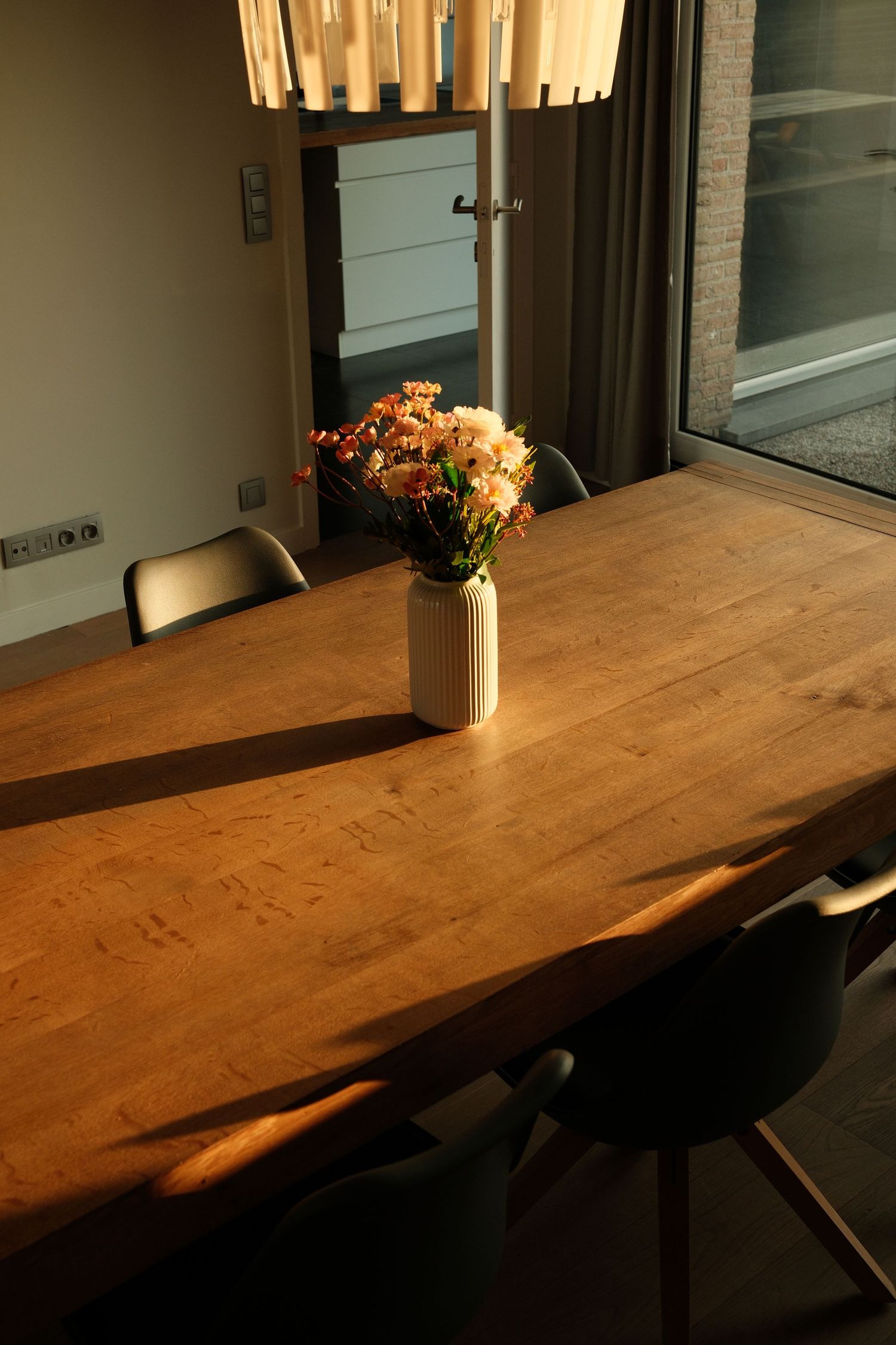 A rustic wooden dining table with dark stain sits near a window with a vase of flowers on top.