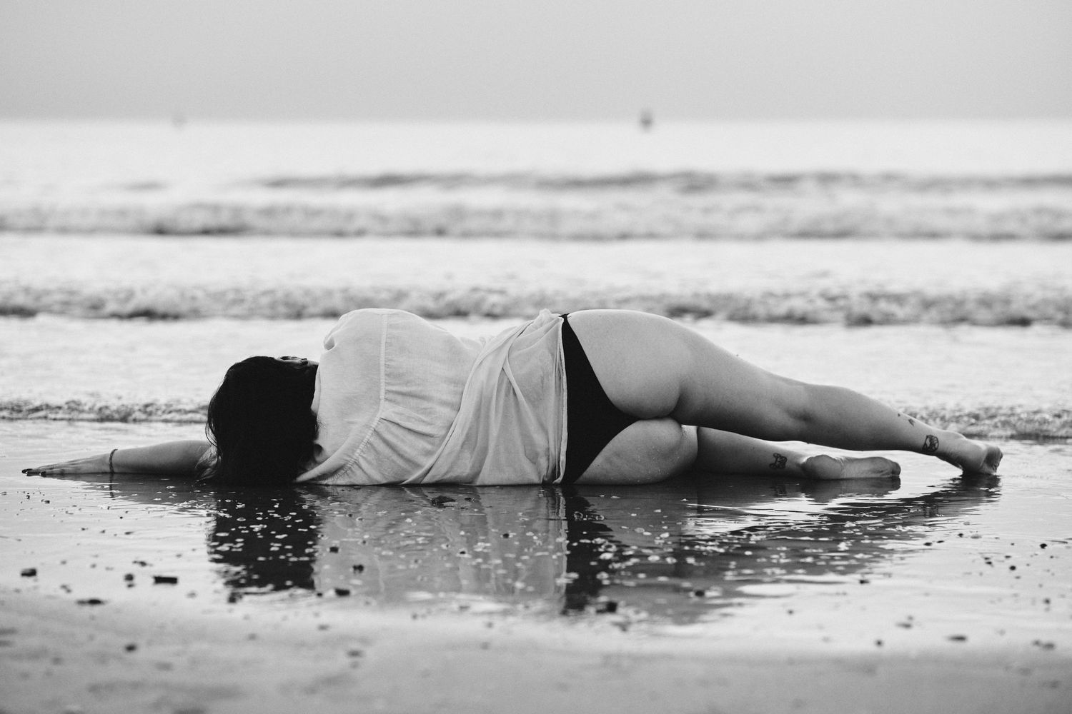 Black and white photo of a woman lying on wet beach sand, draped in white fabric, with ocean waves in the background.
