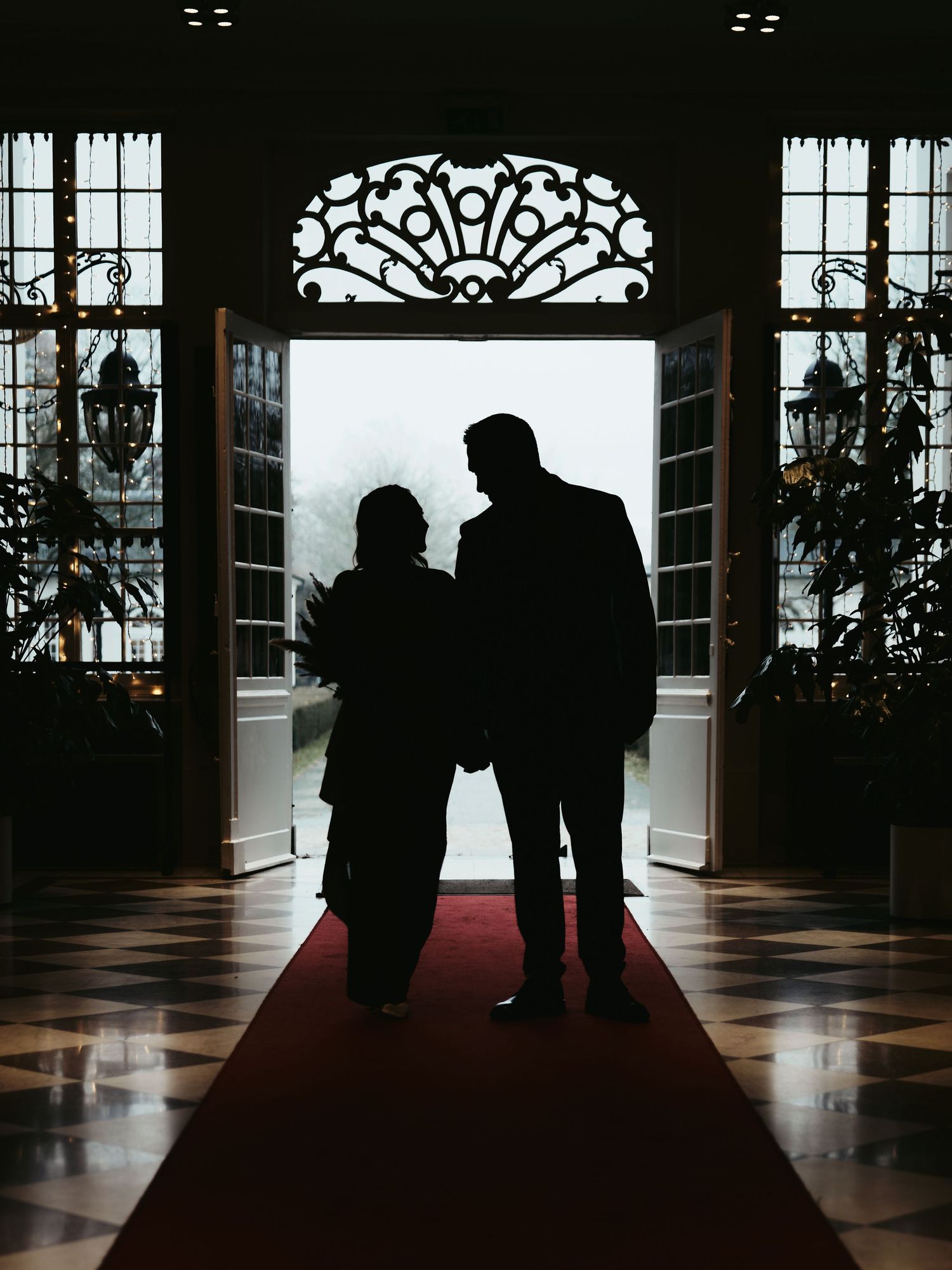 Silhouettes of people standing in a grand hallway with windows and ornate architectural details creating dramatic lighting.
