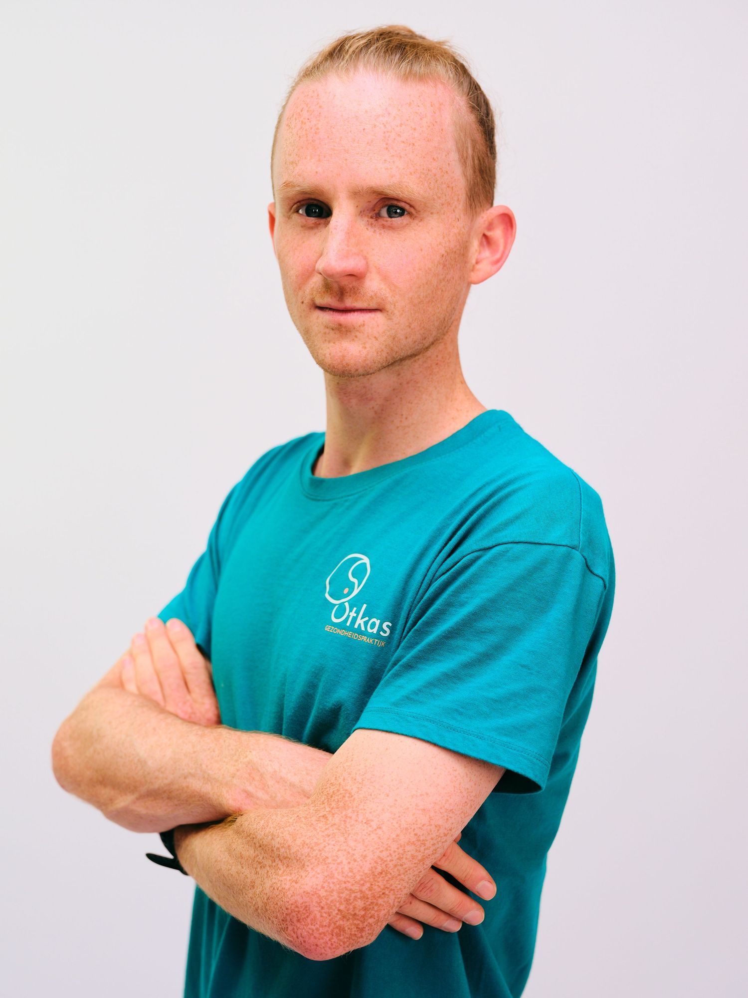 Man in teal Otkas branded t-shirt standing with arms crossed against a light gray background.