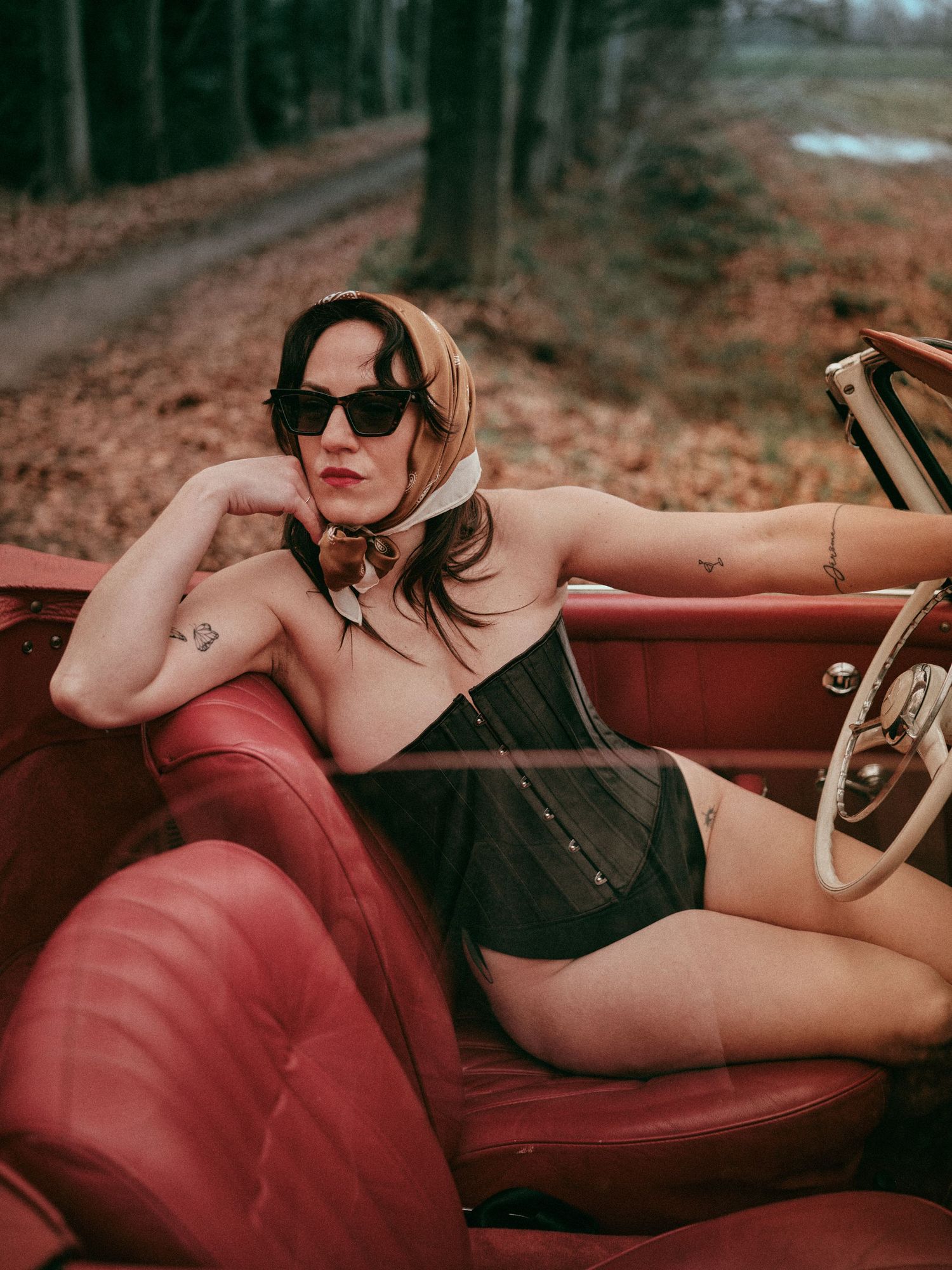 Woman in black dress and sunglasses lounging in vintage red convertible car on a forest road.