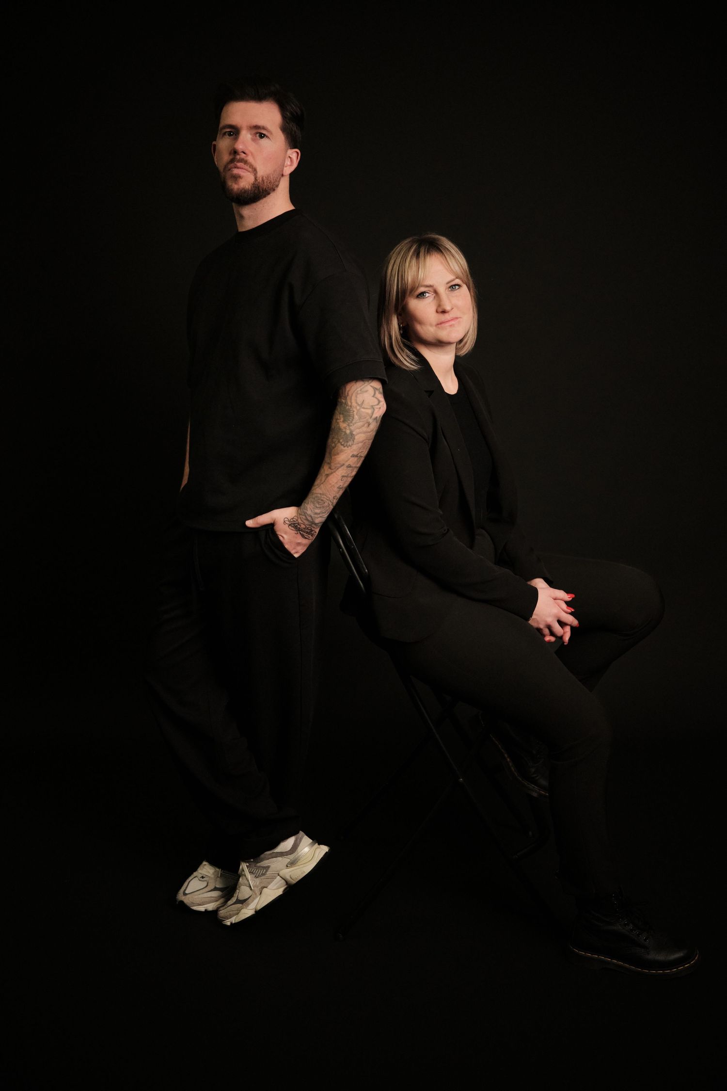 A dramatic studio portrait of two people in black clothing posed against a dark background.