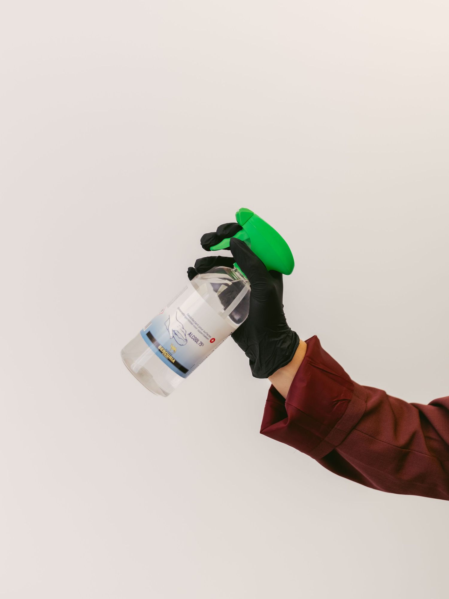 Hand in brown sleeve and green glove holding a small plastic bottle against white background.