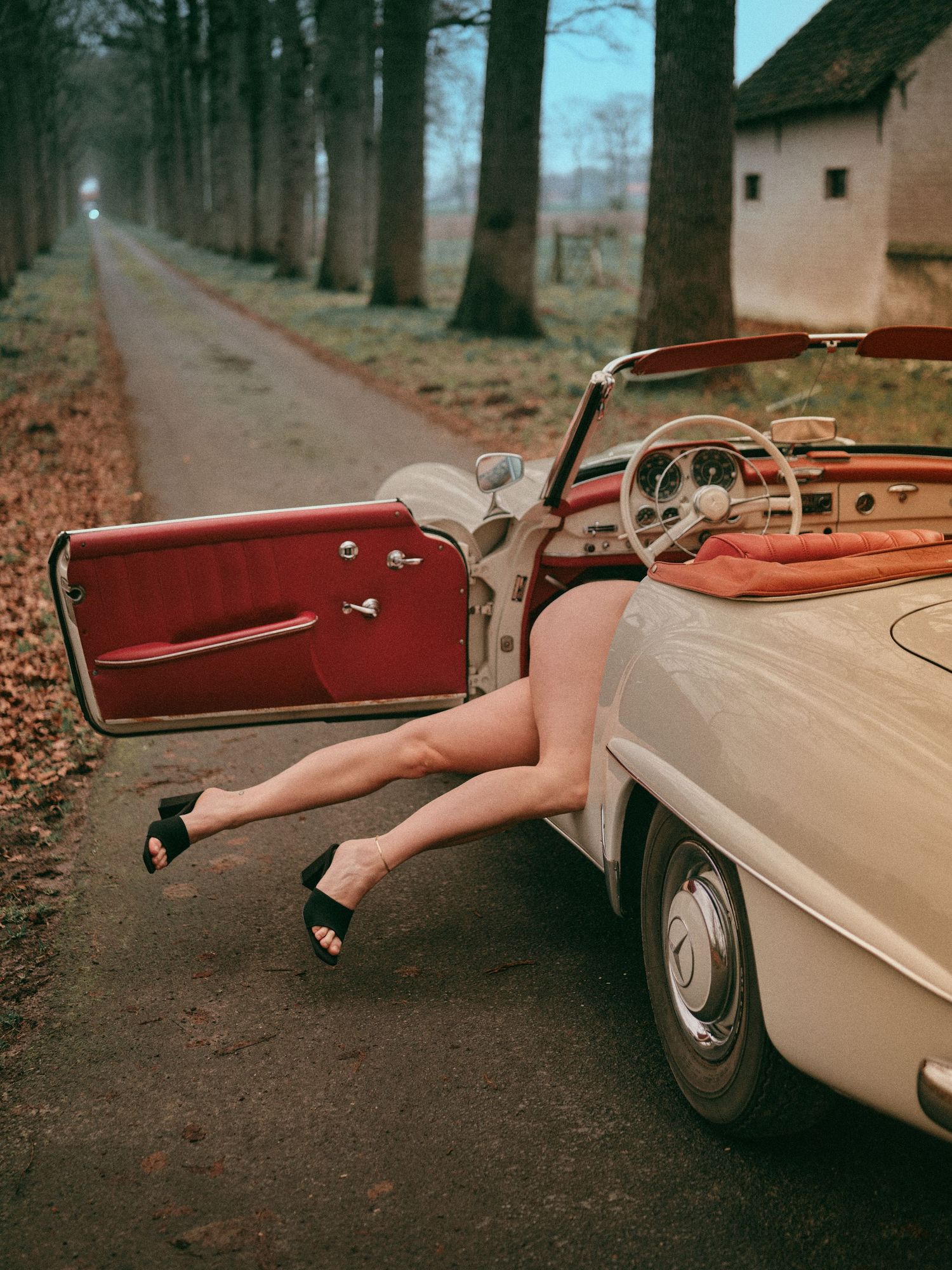 Woman's legs in black heels resting on open door of vintage white Mercedes convertible on a tree-lined road.