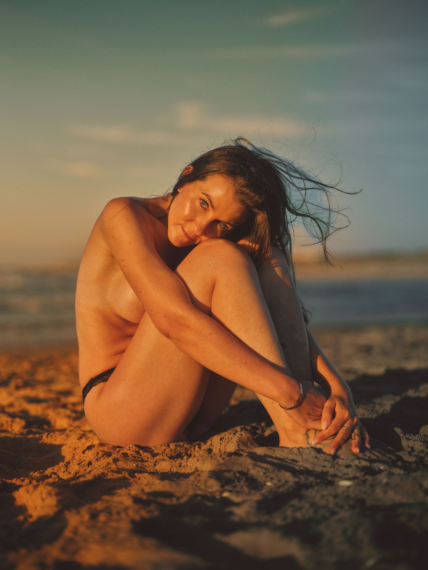 Woman sitting on sandy beach at golden hour sunset, wind blowing hair, ocean in background.