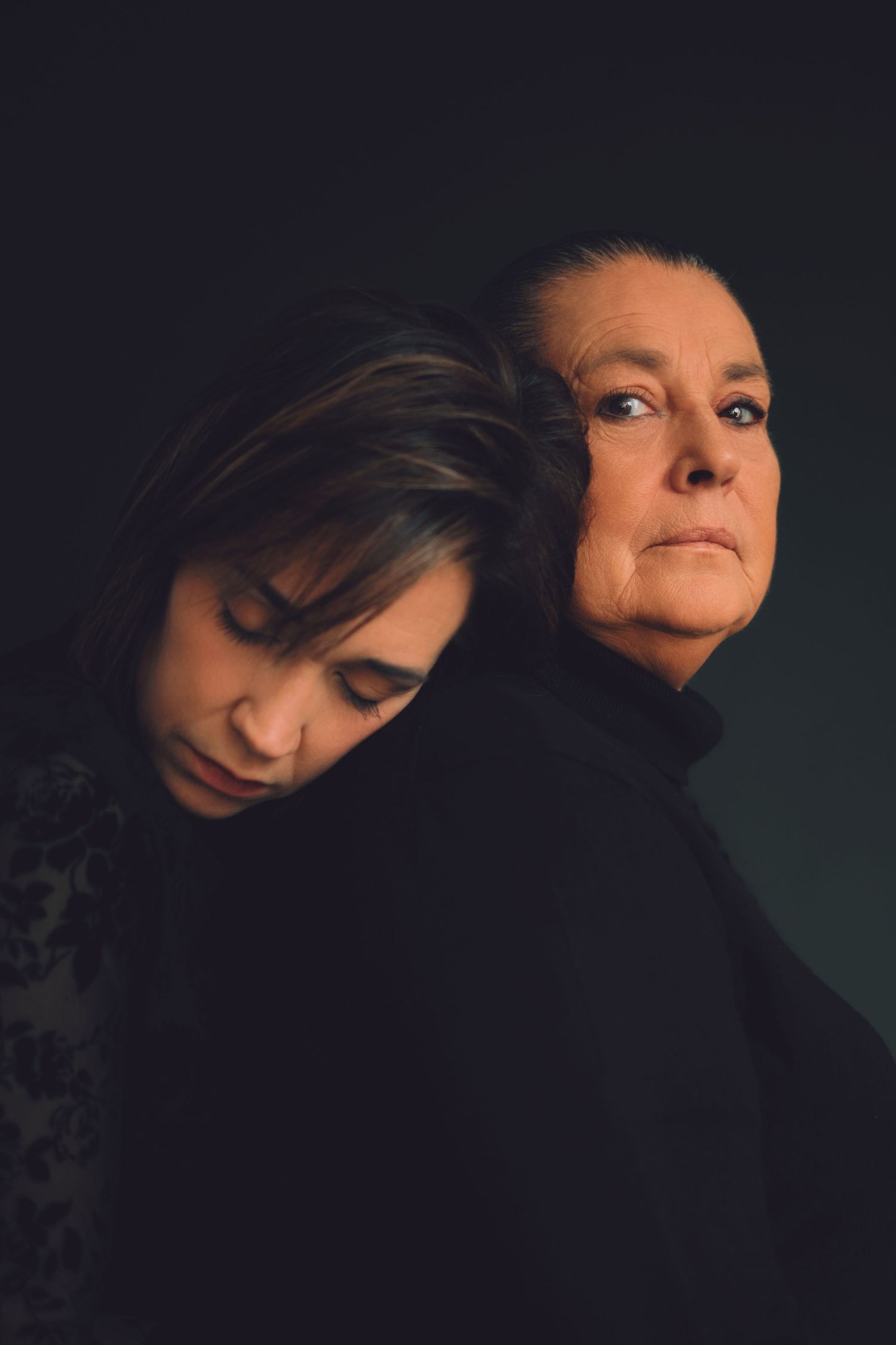 Two women back-to-back in dark setting, younger woman looking down, older woman gazing forward in black turtleneck.