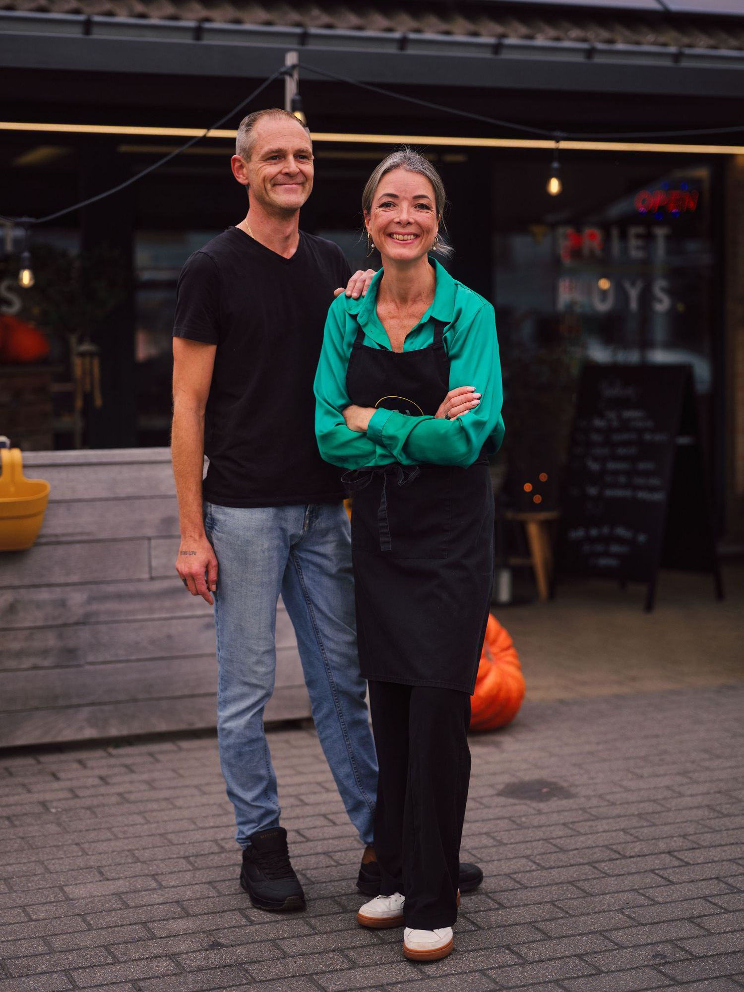 Two smiling restaurant owners pose outside their eatery, woman in green shirt and black apron, man in black t-shirt and jeans.