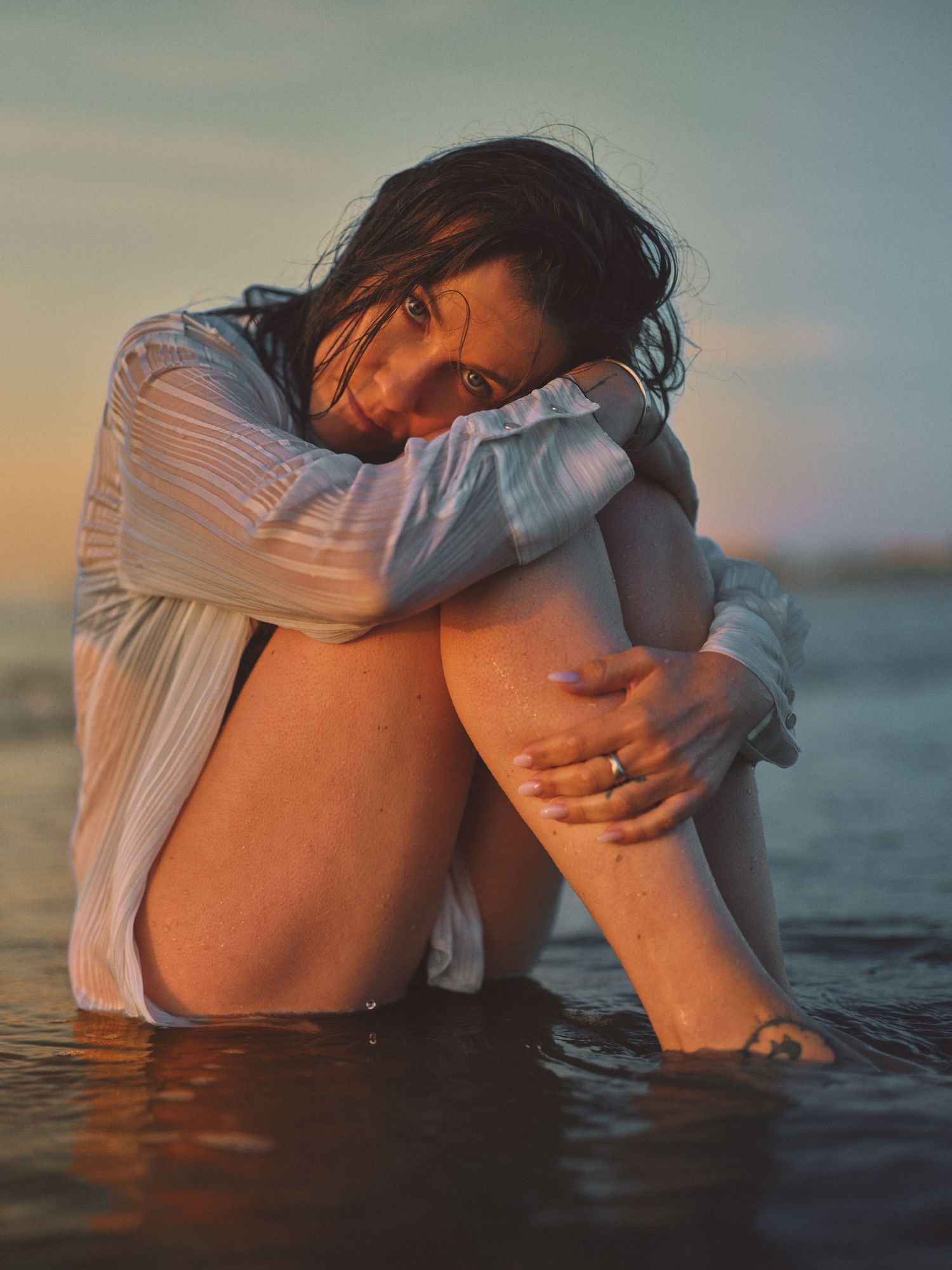 Woman in wet white shirt sitting in shallow water at sunset, hugging her knees in a contemplative pose.