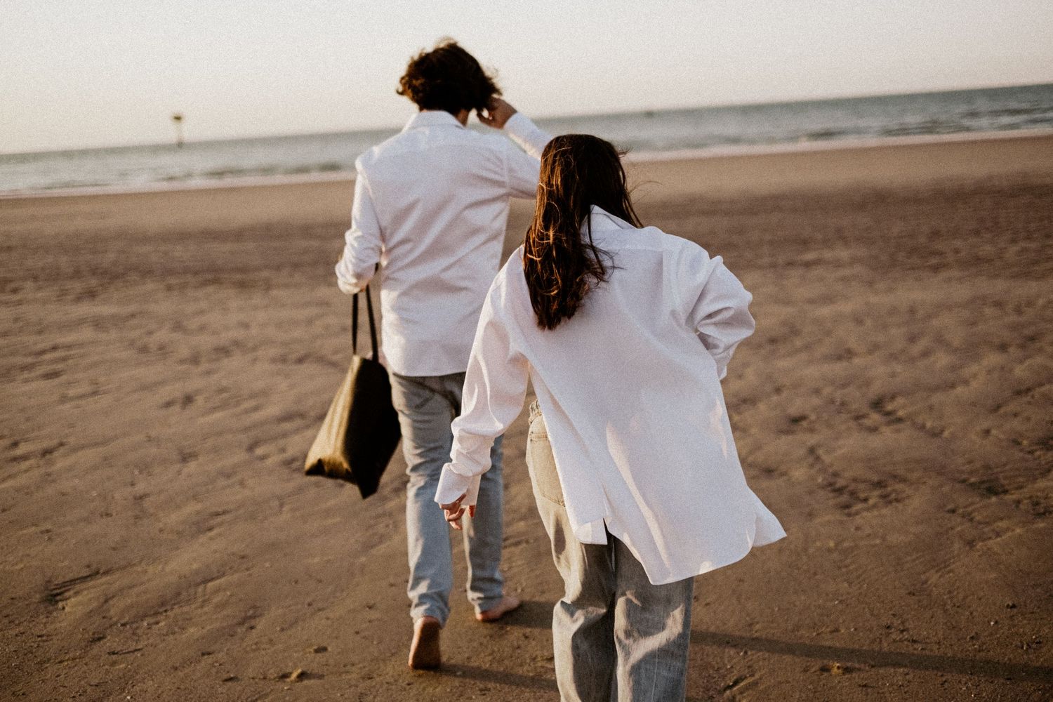 Two people in white shirts and jeans walk hand in hand on a sandy beach at sunset.