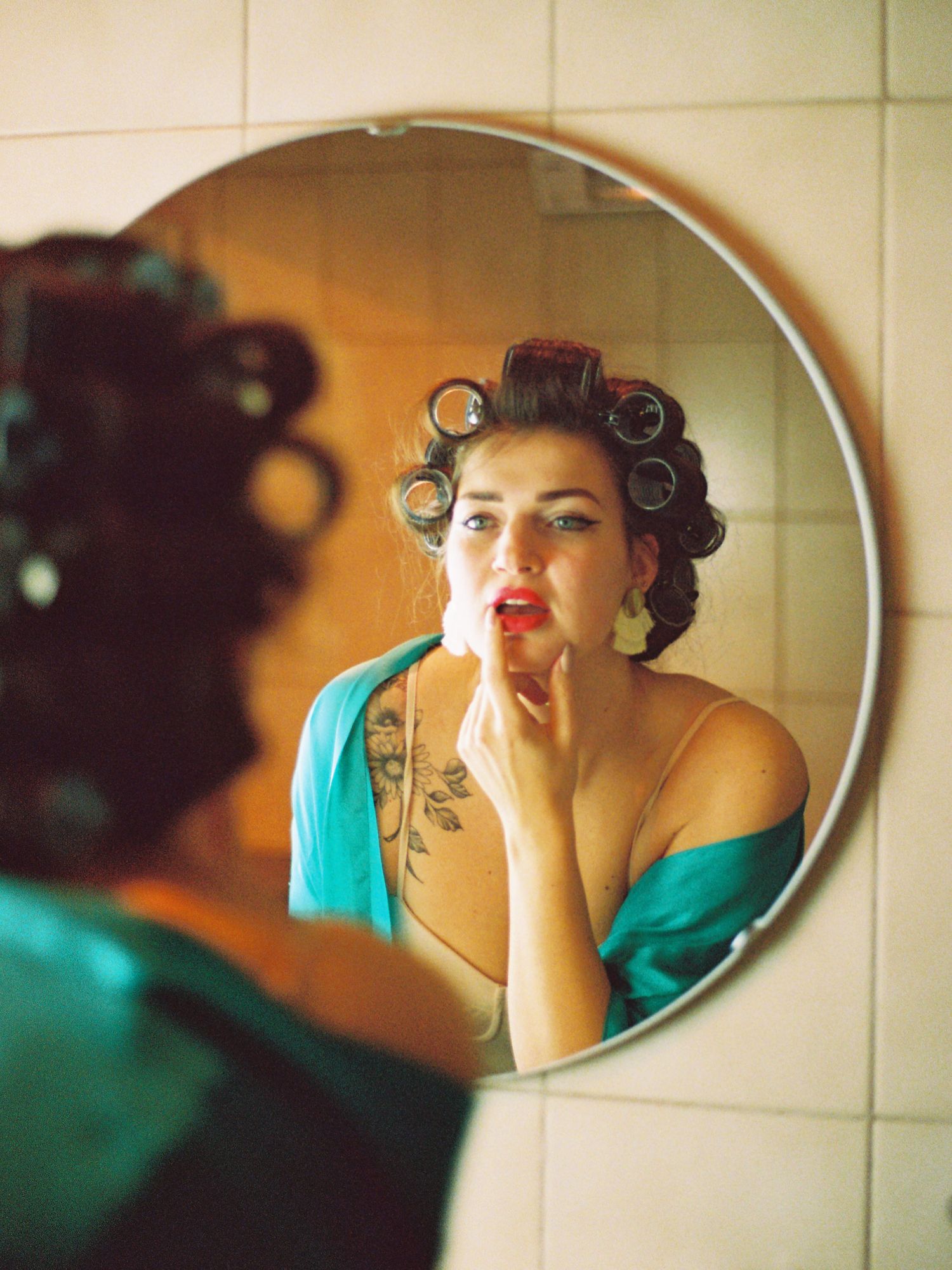 Woman with hair rollers applying red lipstick while looking in a round bathroom mirror, wearing a teal robe.
