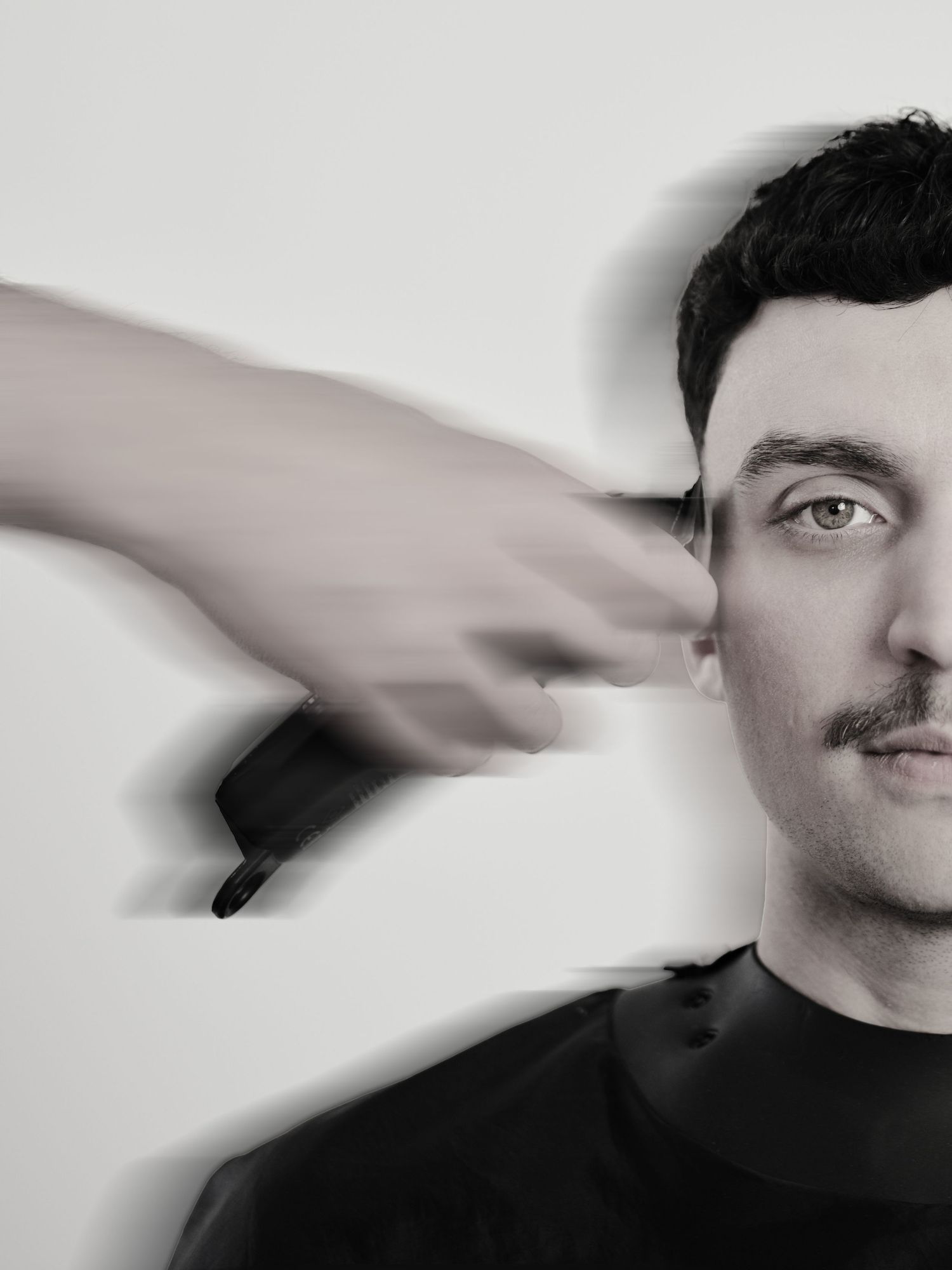 Black and white photo of a man with mustache getting hair trimmed with clippers at a barbershop.