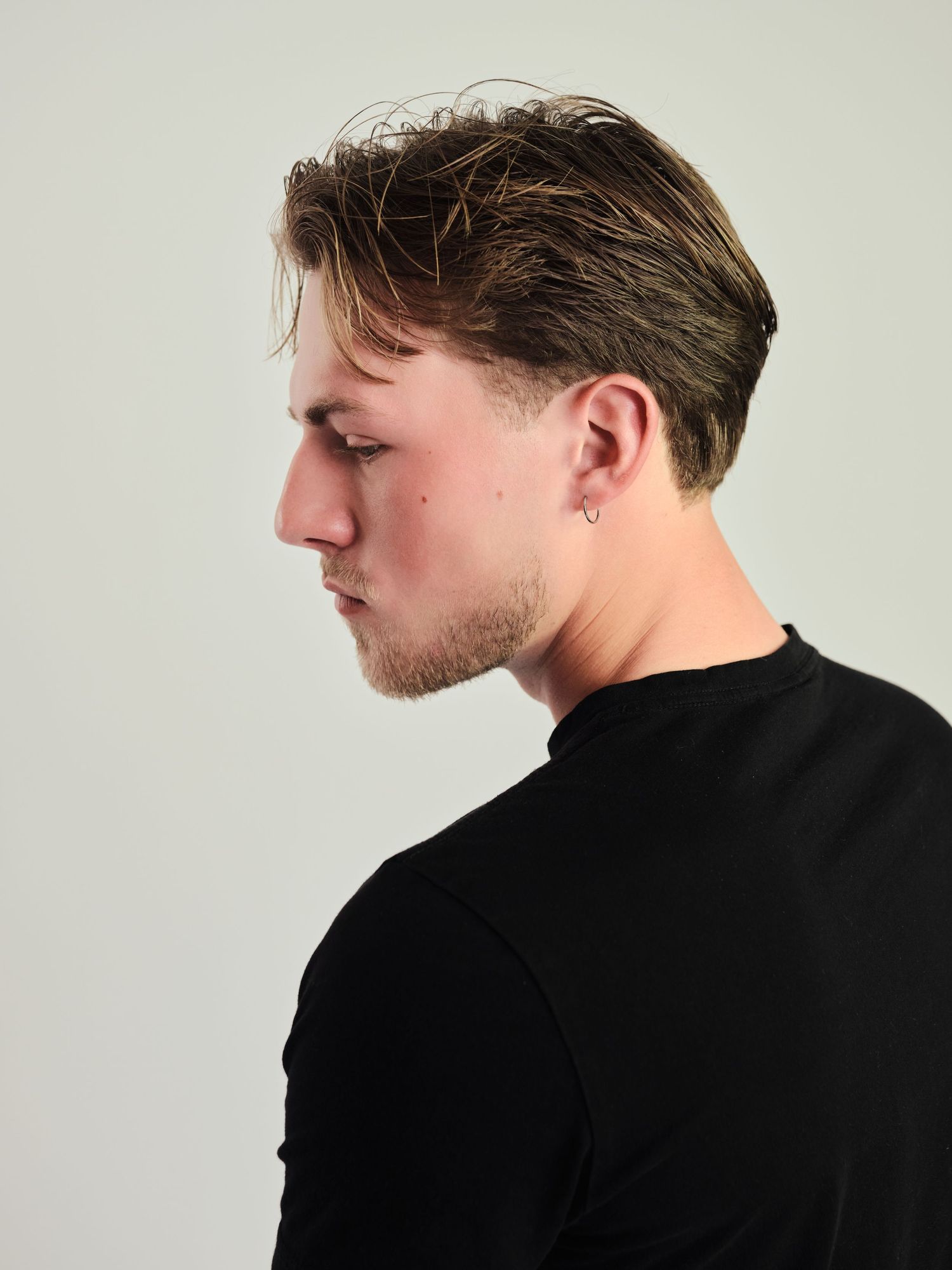 Young man in profile view wearing black turtleneck, short brown hair, small hoop earring, light stubble, white background.
