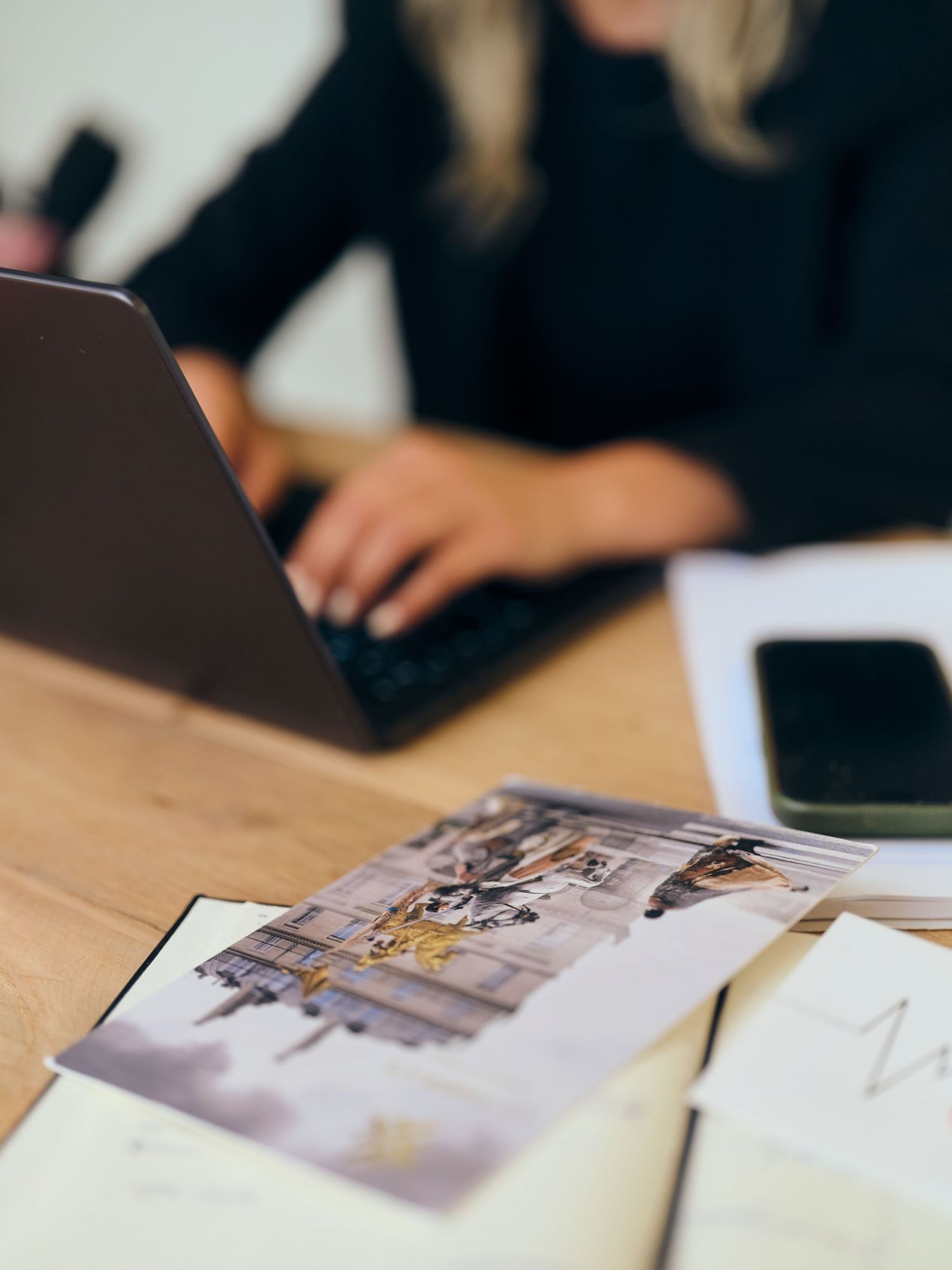 Woman working on laptop at wooden desk with printed photos, smartphone, and papers in a modern office setting.