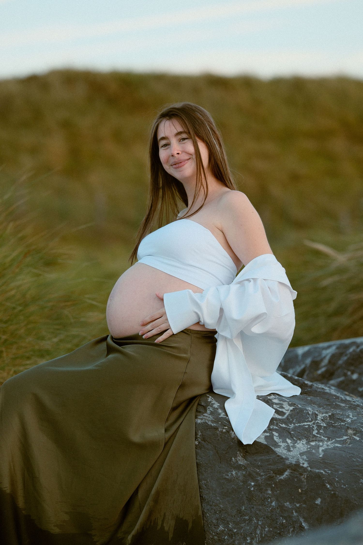 Artistic maternity portraits in a natural setting with flowing white dress against grassy backdrop.