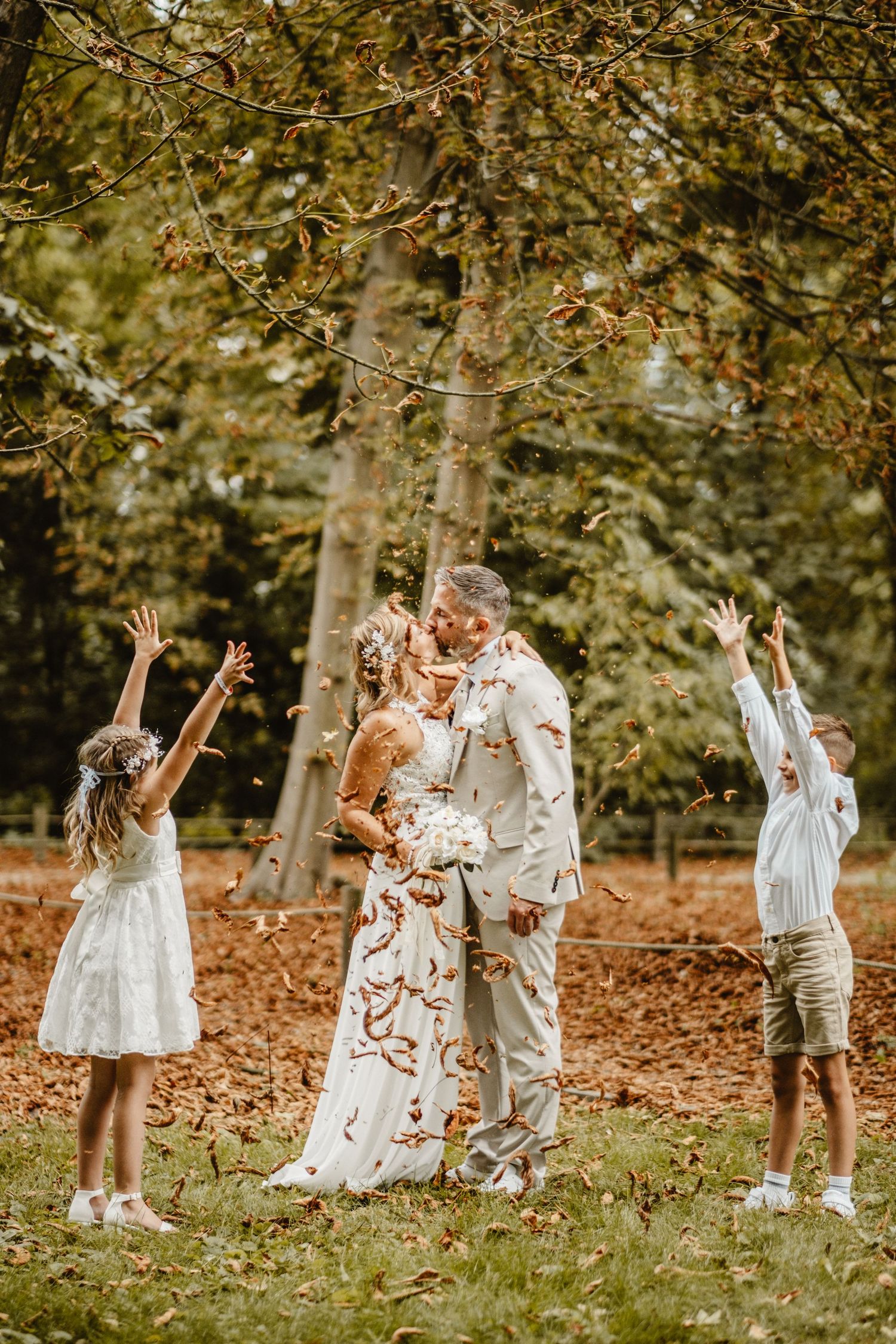 Wedding party throws autumn leaves in celebration outdoors among trees.