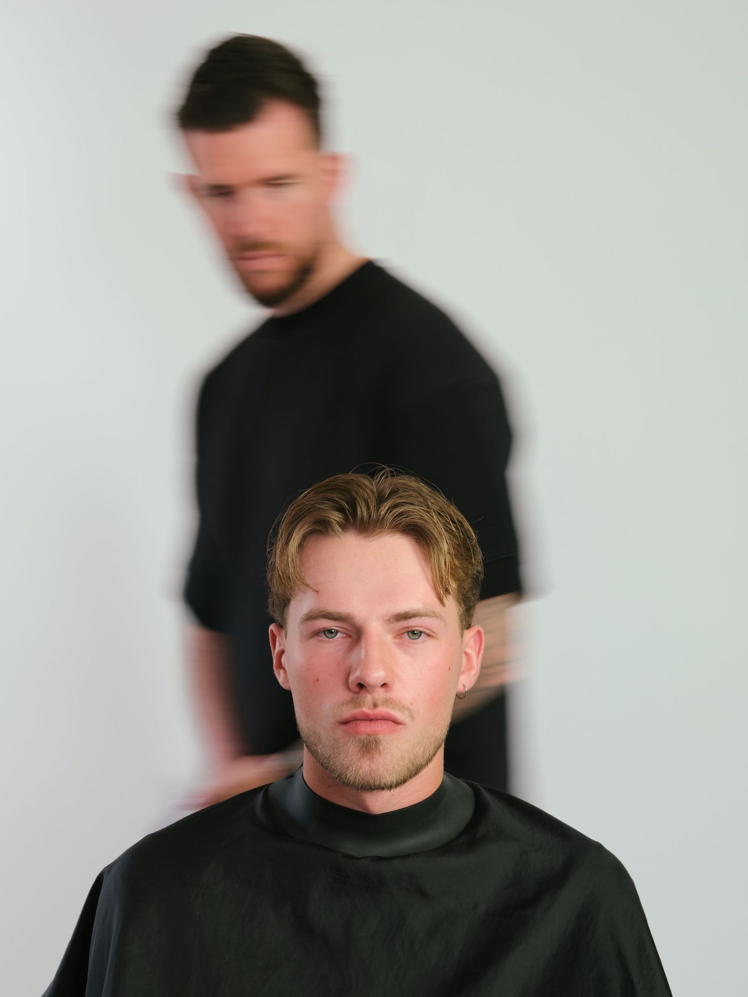 Man in barber cape getting haircut with blurred motion of barber in black t-shirt behind him.