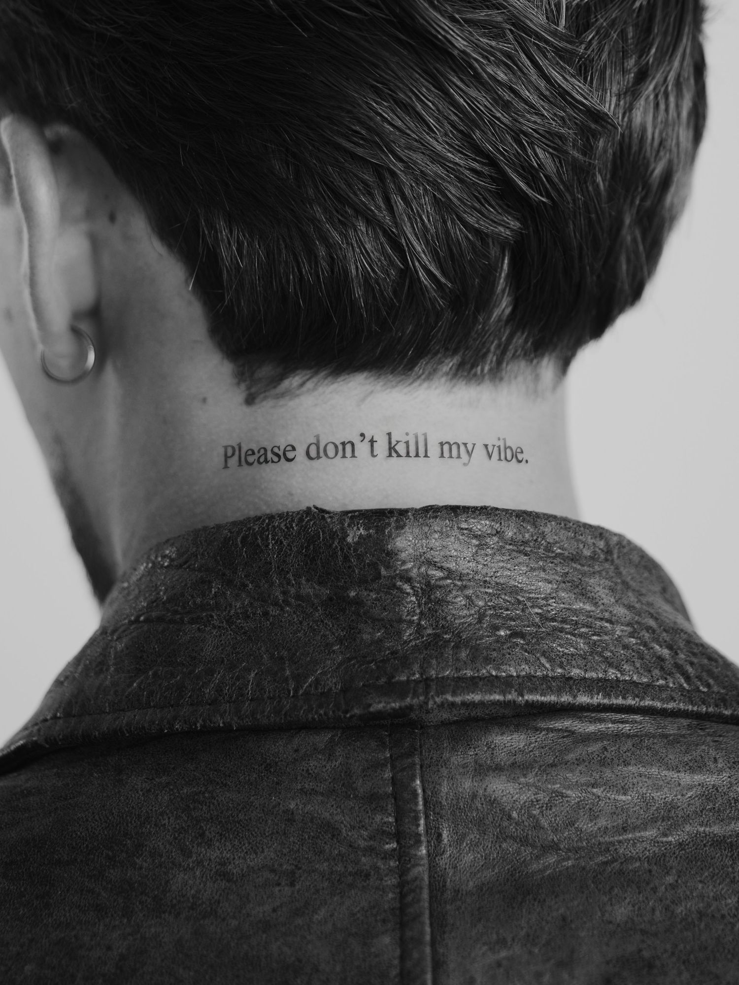 Black and white photo of neck tattoo reading Please don't kill my vibe on person wearing leather jacket.