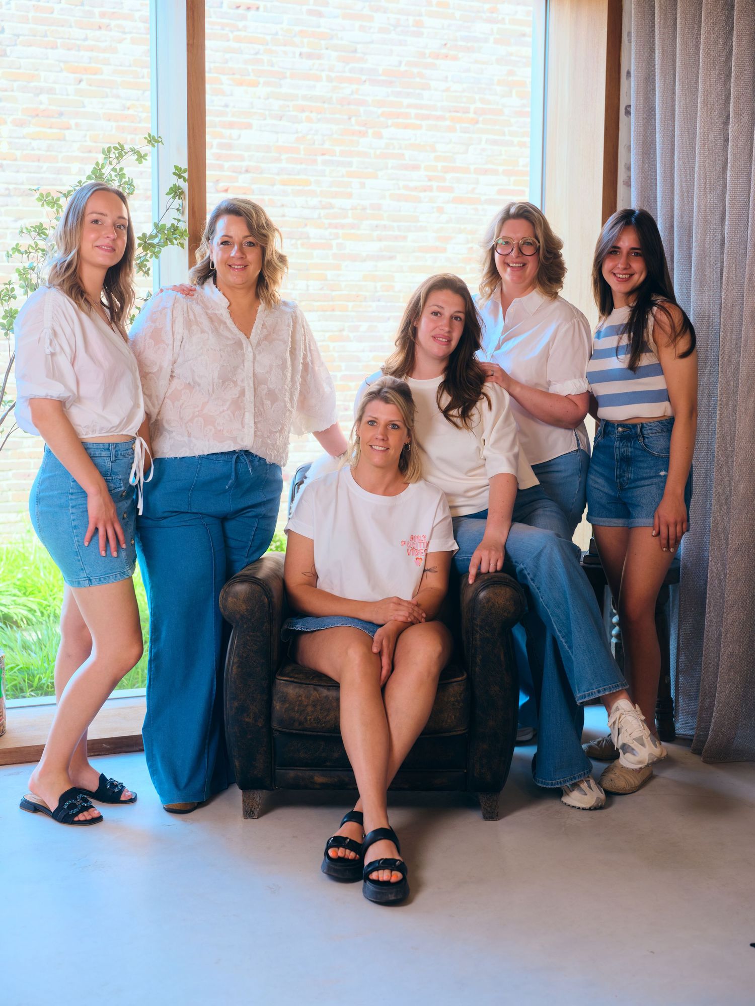 Six women posing together indoors, five standing and one seated in a leather chair, wearing casual blue and white outfits.