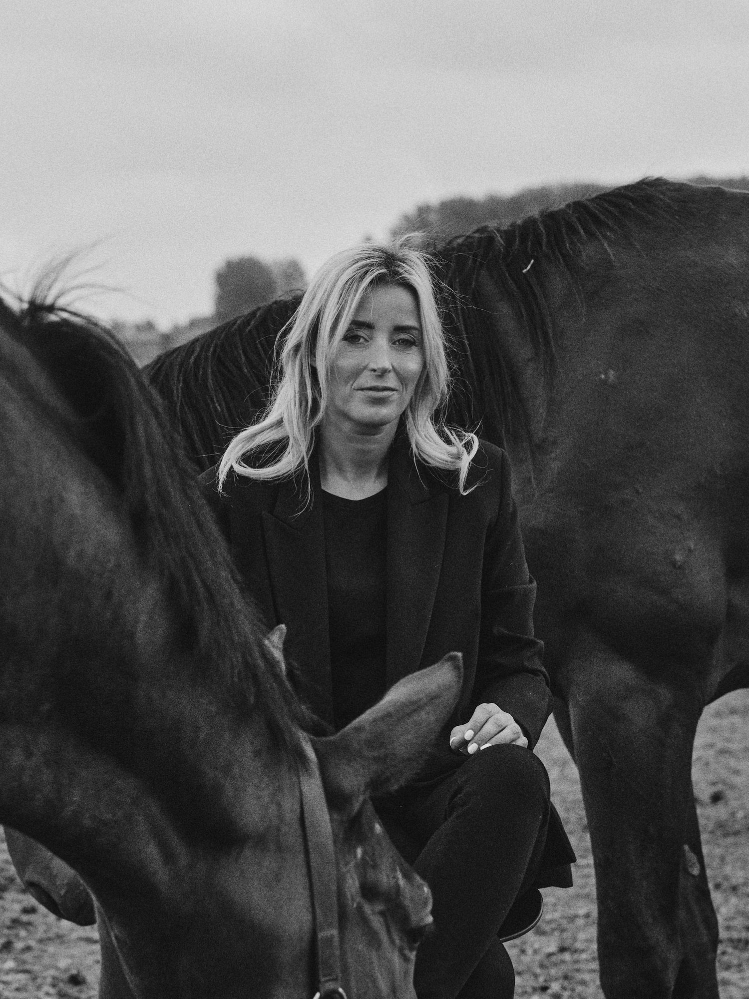 Black and white photo of a blonde woman in a black outfit crouching between two dark horses outdoors.