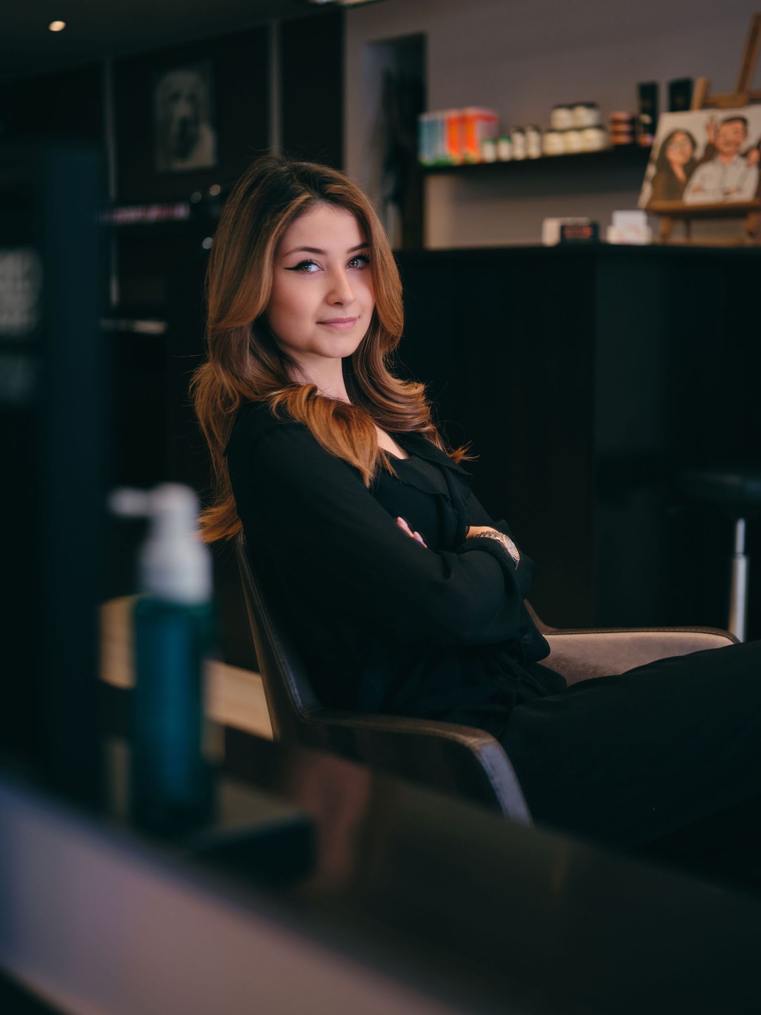 Woman in black outfit sits confidently in a modern salon chair with arms crossed, surrounded by beauty products.