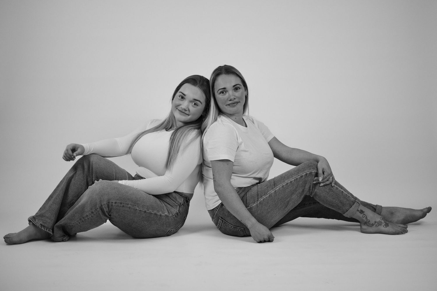 Two young women sitting back-to-back on floor, smiling, wearing white tops and jeans, black and white studio photo.
