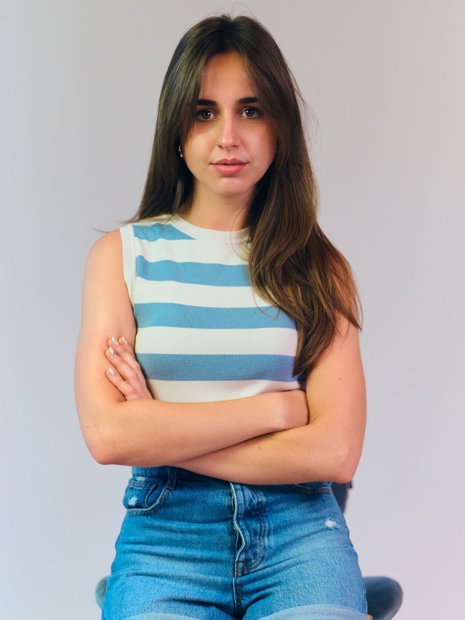 Young woman with long brown hair wearing a striped blue and white sleeveless top and jeans, arms crossed.