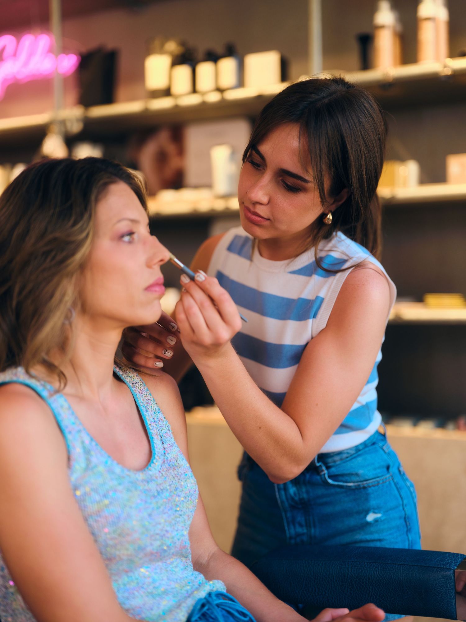 A makeup artist applies foundation with a brush to a woman in a sequin top at a beauty salon.