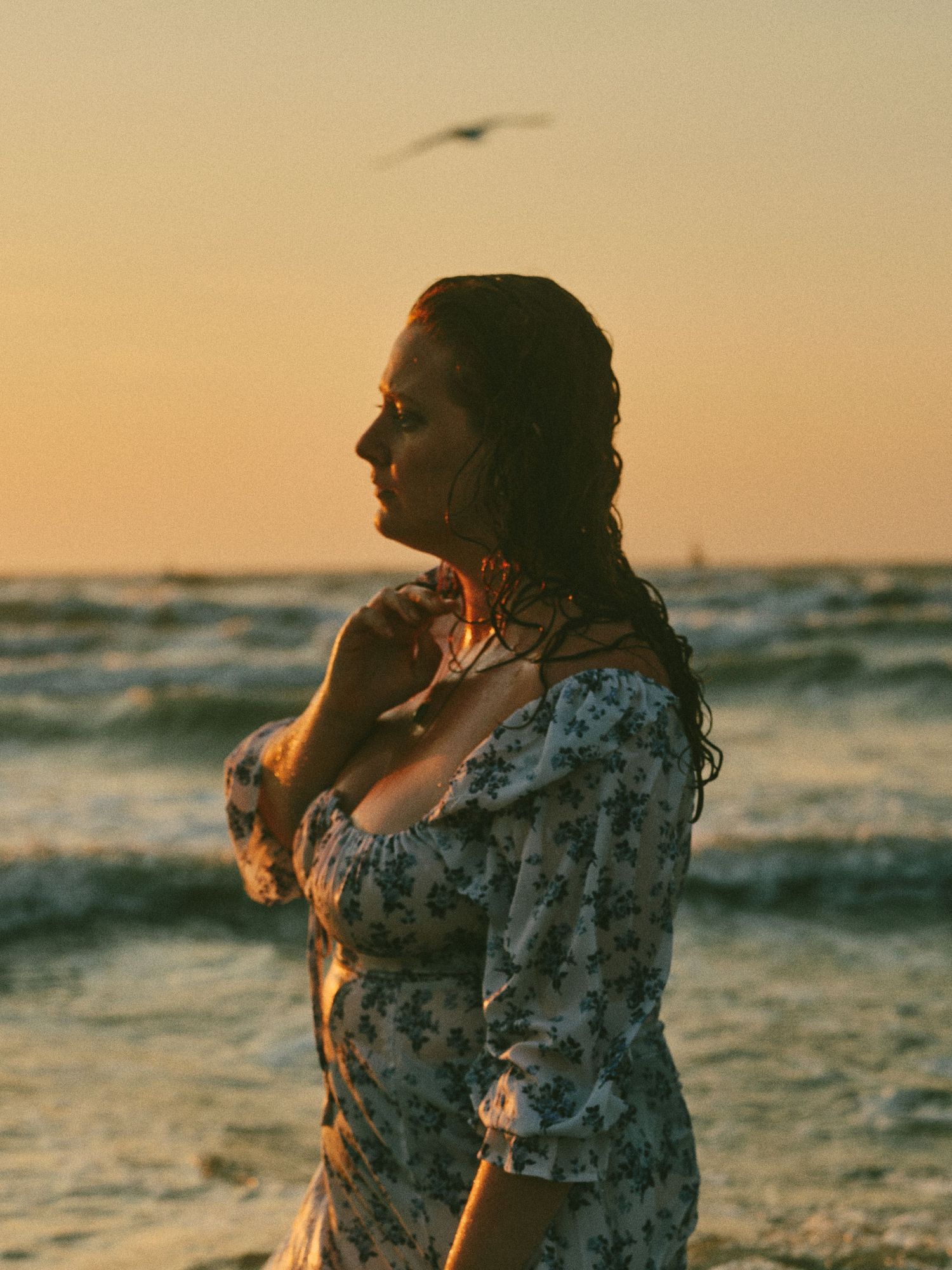 Woman in floral dress standing on beach at golden sunset with ocean waves and seagull in background.