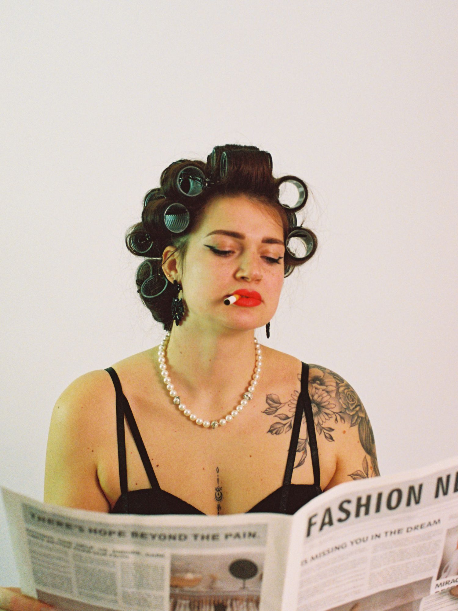 Woman with hair rollers, red lipstick, pearl necklace, and tattoos reading a fashion newspaper.