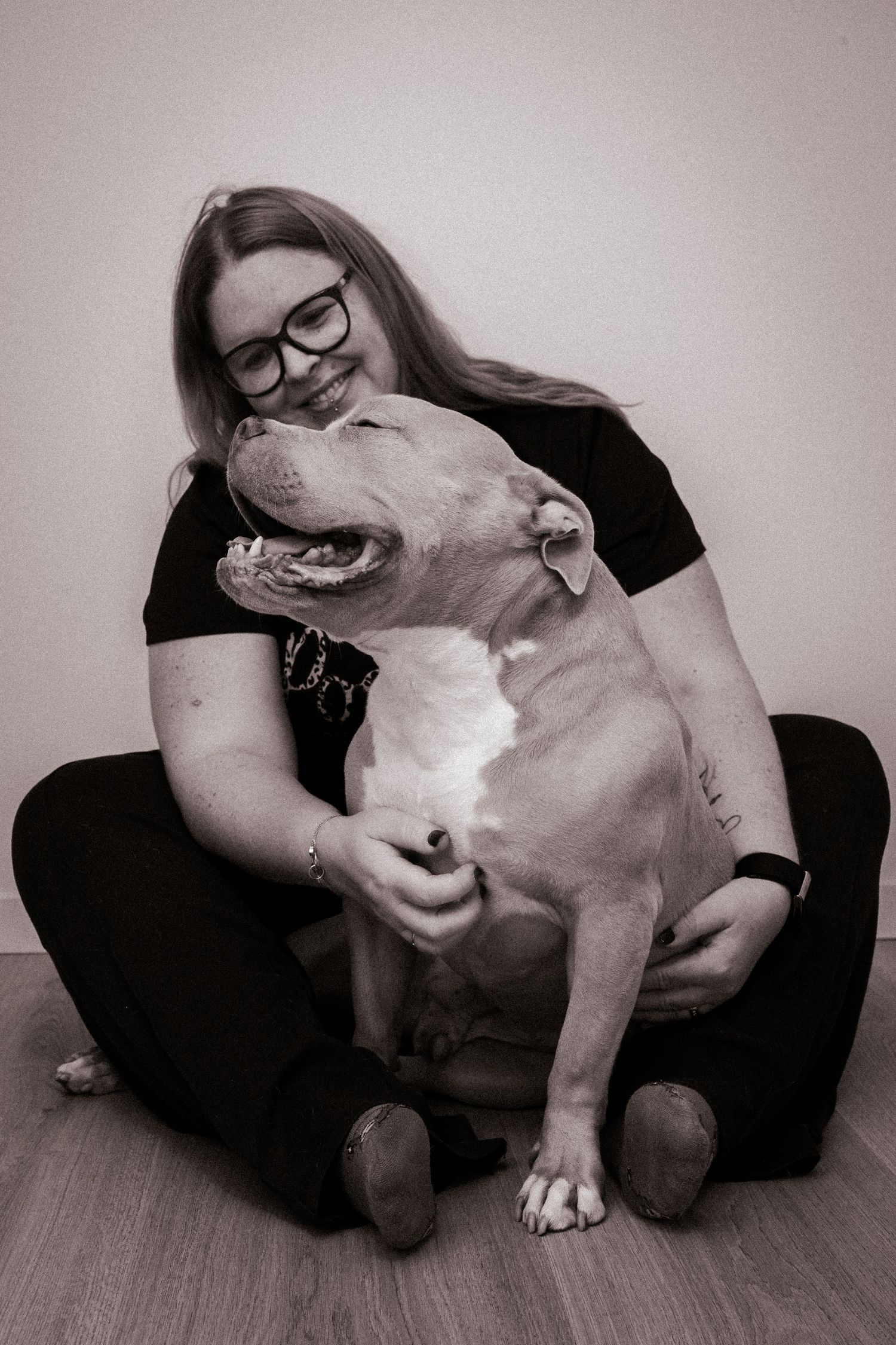 A happy pit bull dog sitting on the floor with someone in a black shirt in a black and white photograph.