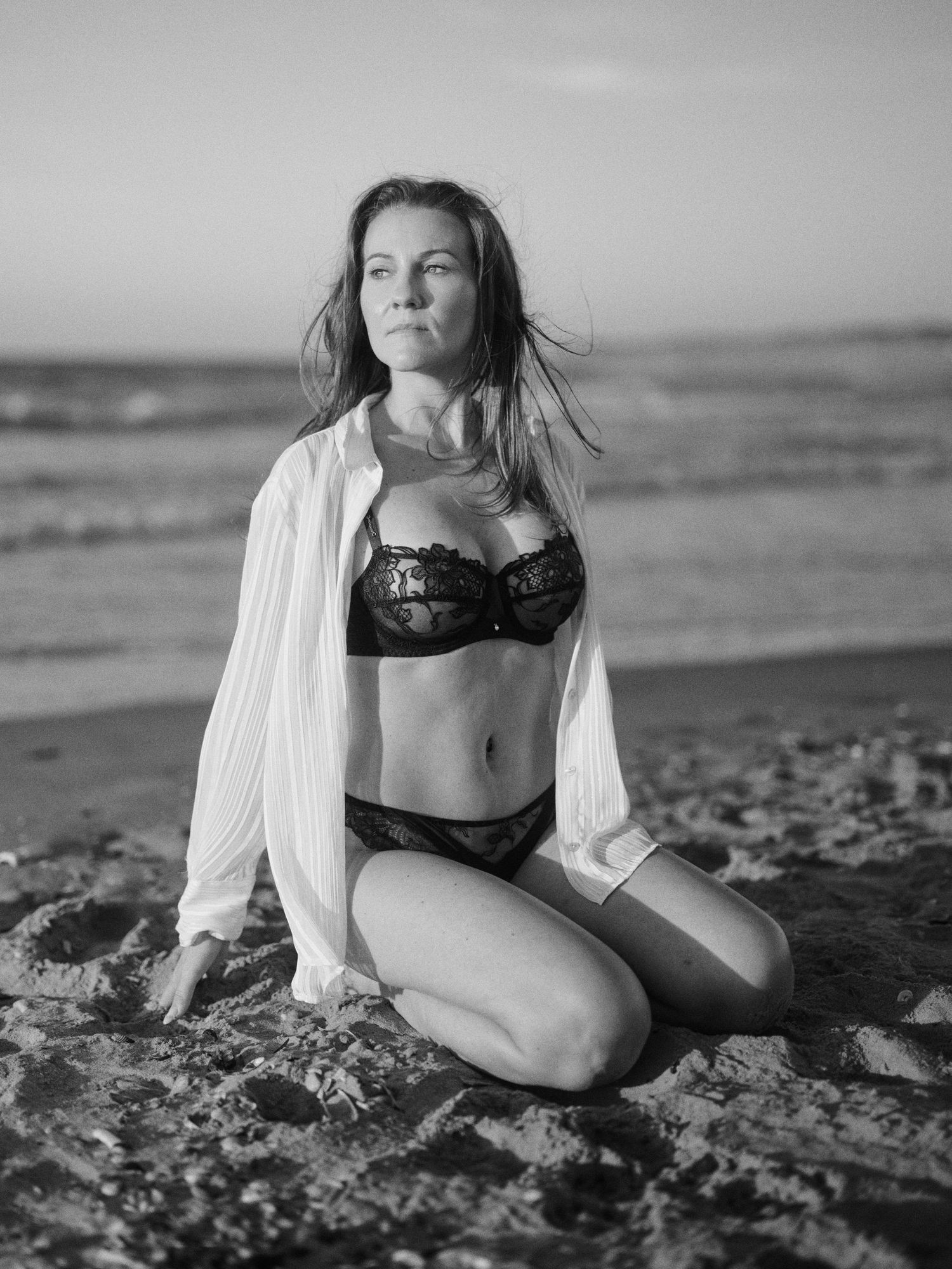 Black and white photo of a woman in lace lingerie and open shirt kneeling on a sandy beach with ocean waves behind her.