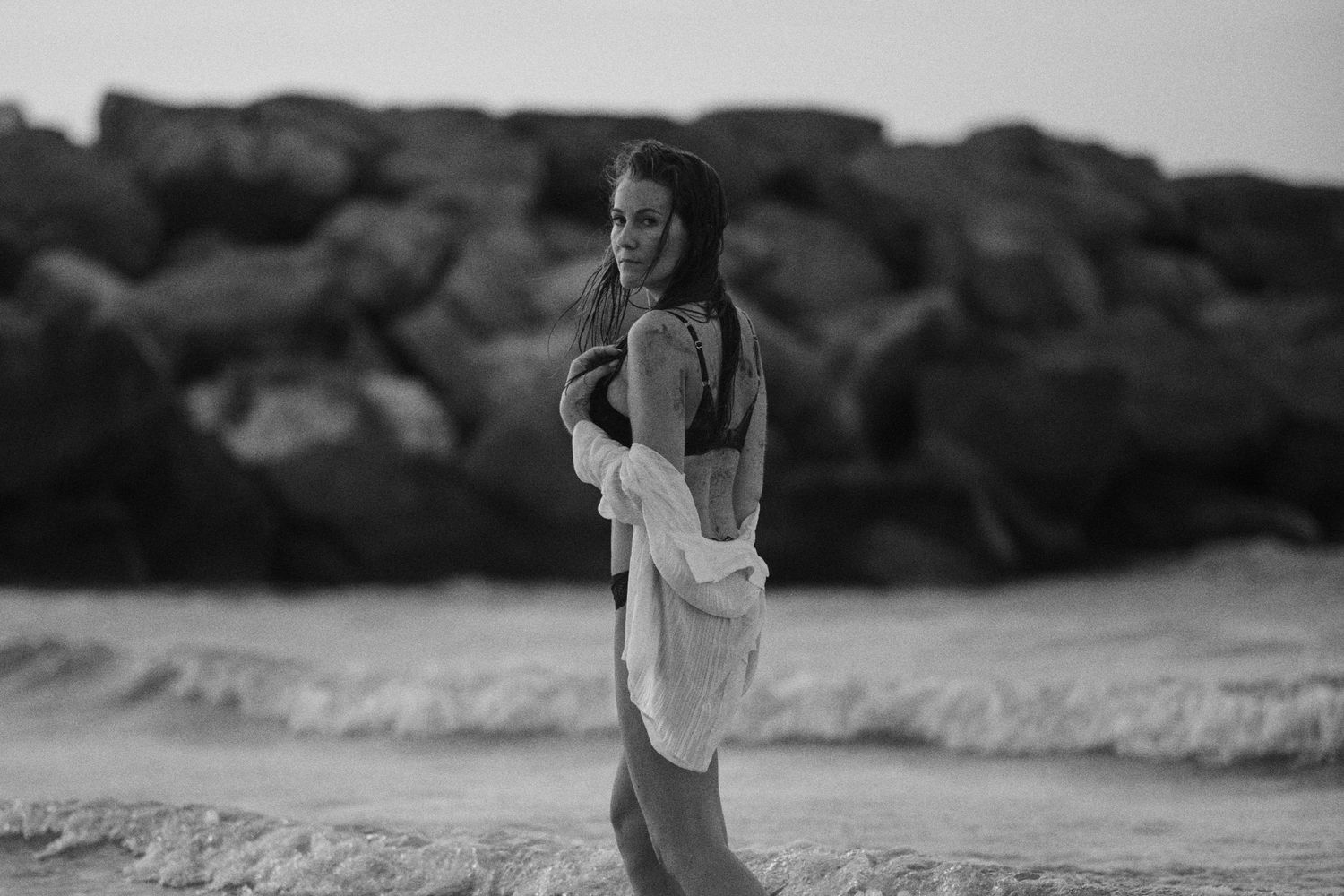 Black and white photo of a woman in a white shirt posing on a rocky beach with ocean waves behind her.