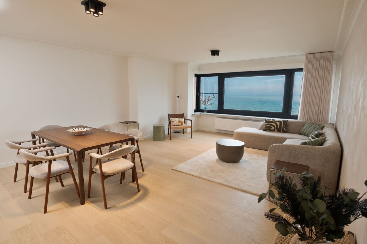 Modern living and dining room with ocean view, featuring a beige sofa, wooden dining set, and light hardwood floors.