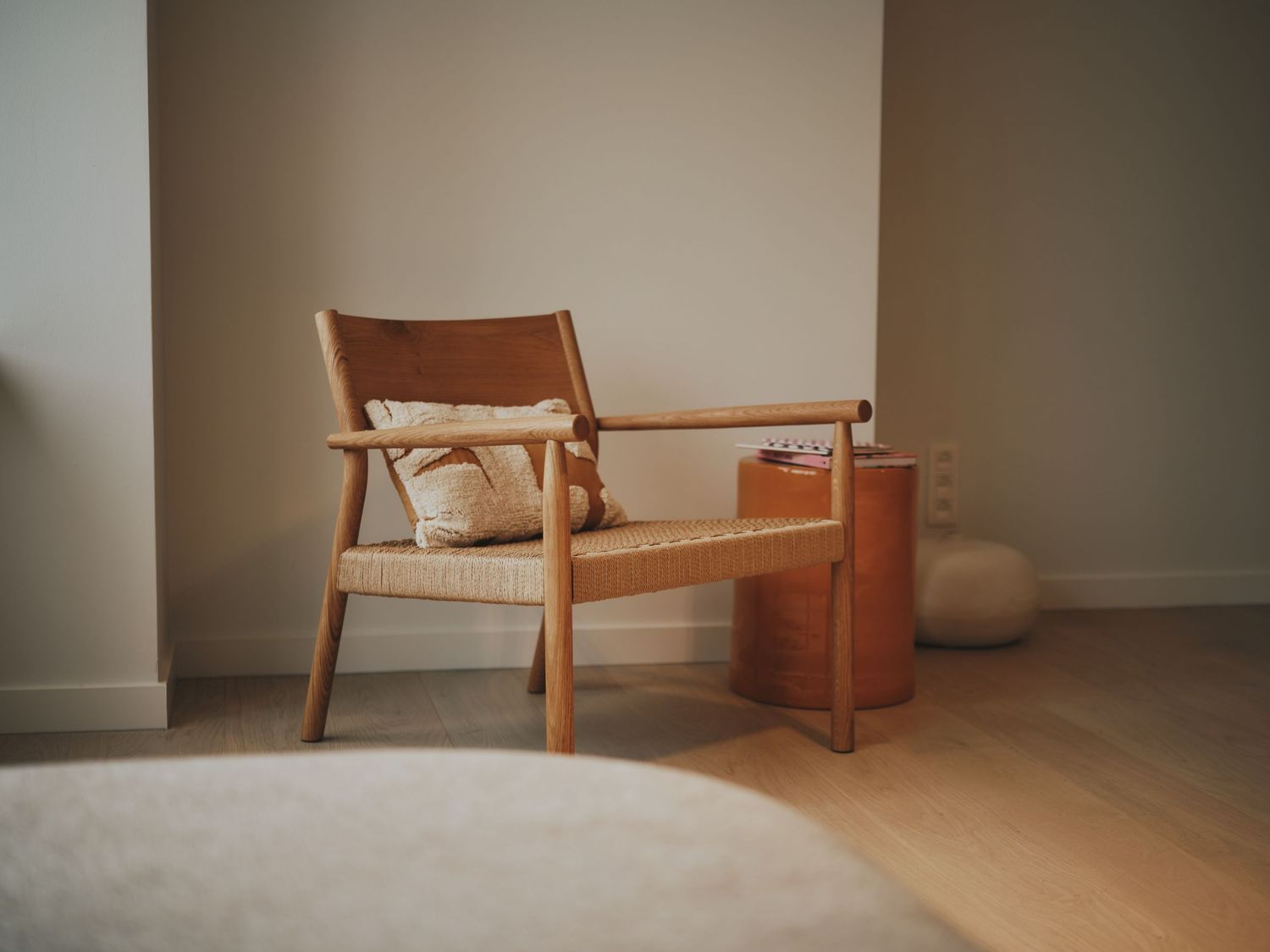 Minimalist living room with a wooden lounge chair, woven seat cushion, and small side table on light hardwood floors.