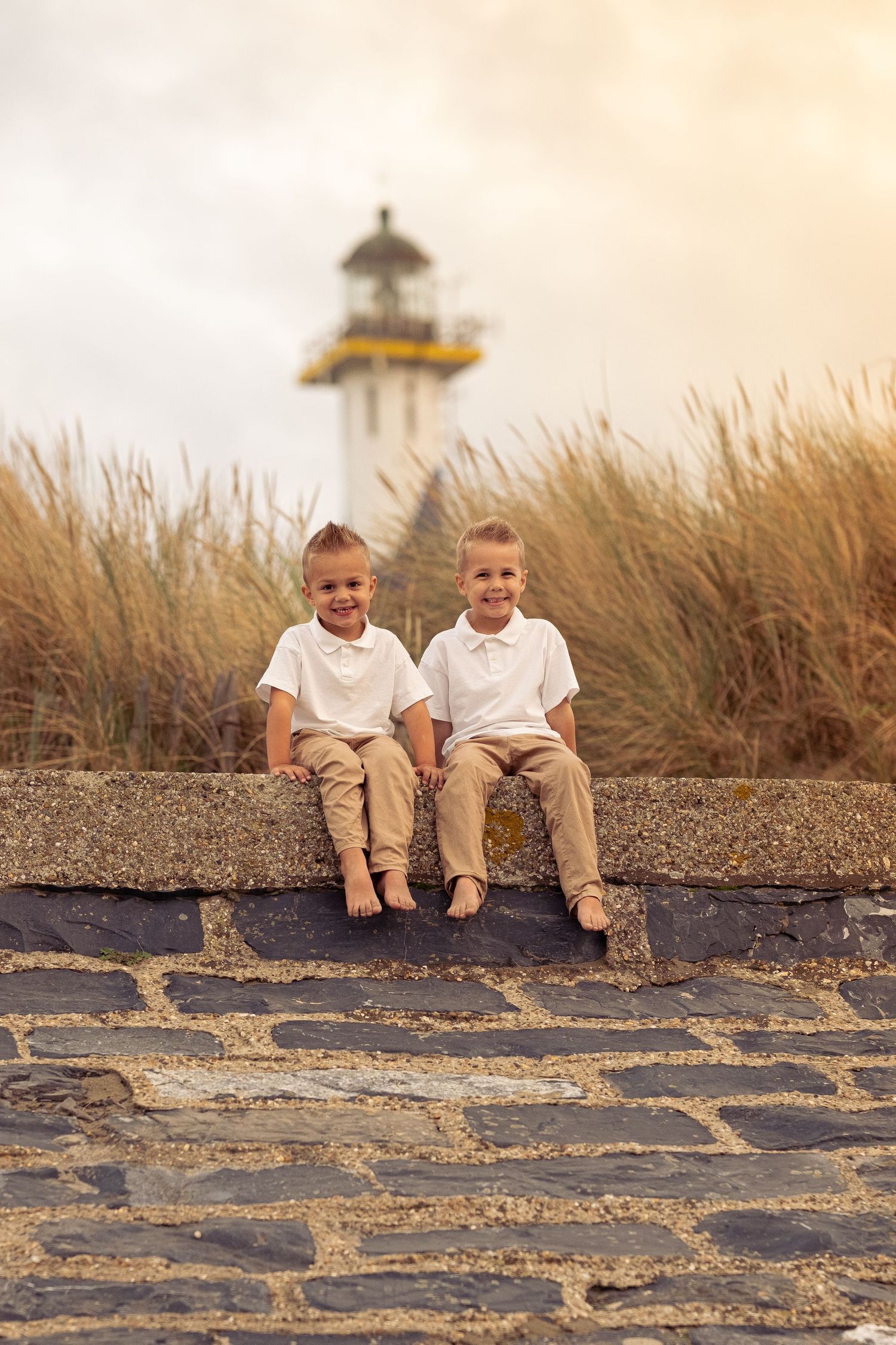 Children sitting on stone wall with lighthouse and coastal scenery in background.