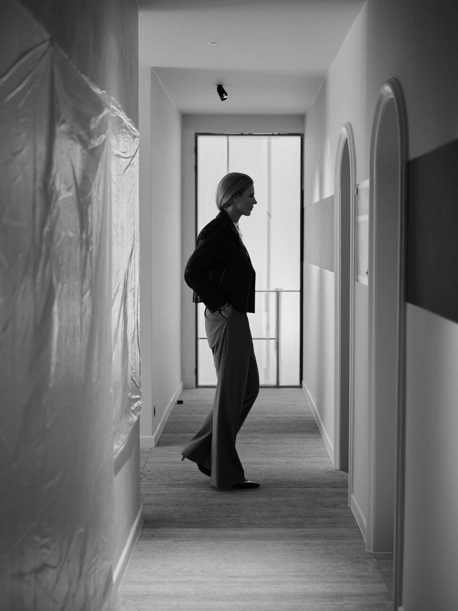 Black and white photo of a person walking down a narrow hallway toward a window, wearing dark jacket and wide-leg trousers.