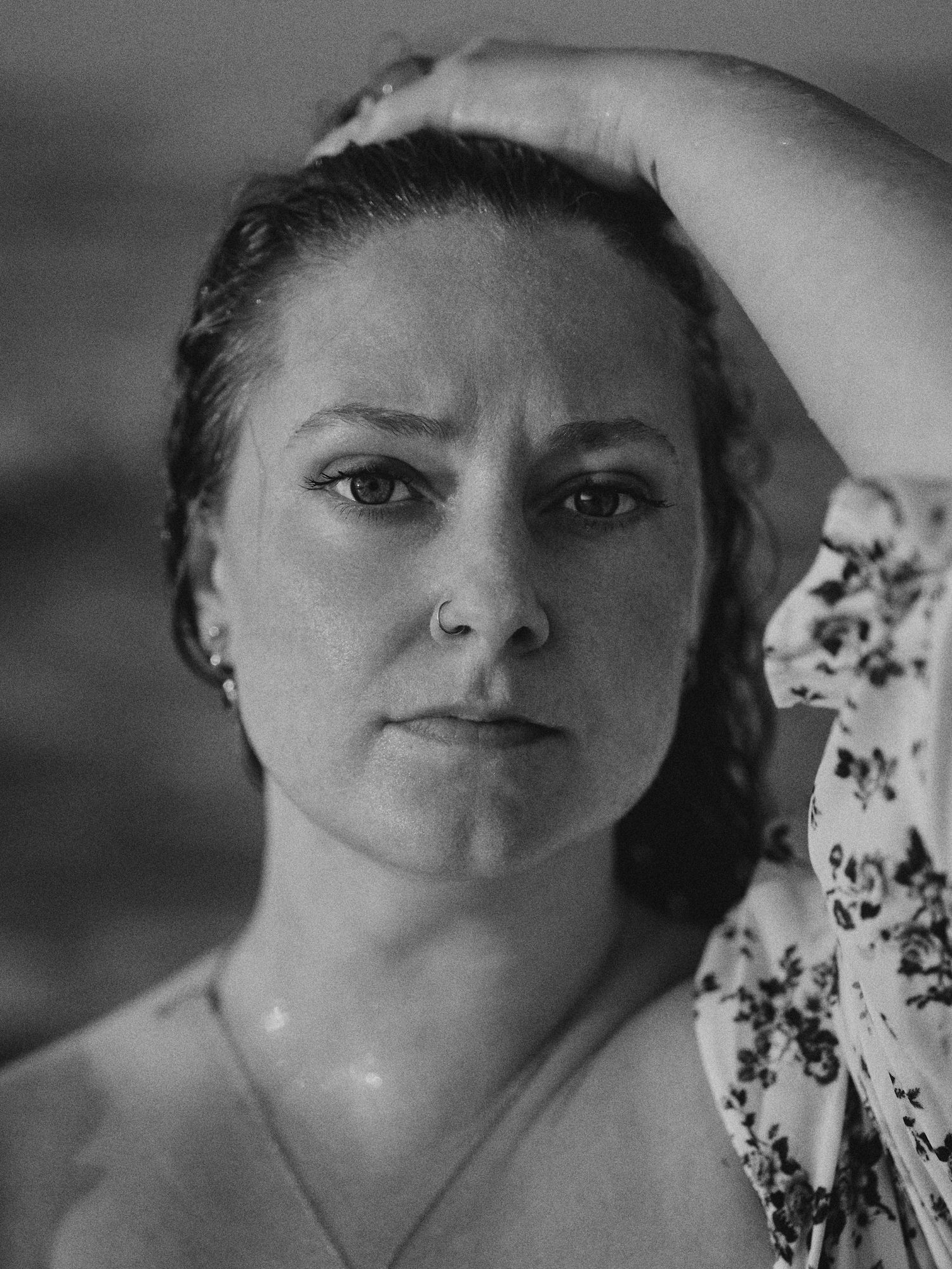 Black and white portrait of a woman with wet hair, nose piercing, and floral top, hand raised to head.
