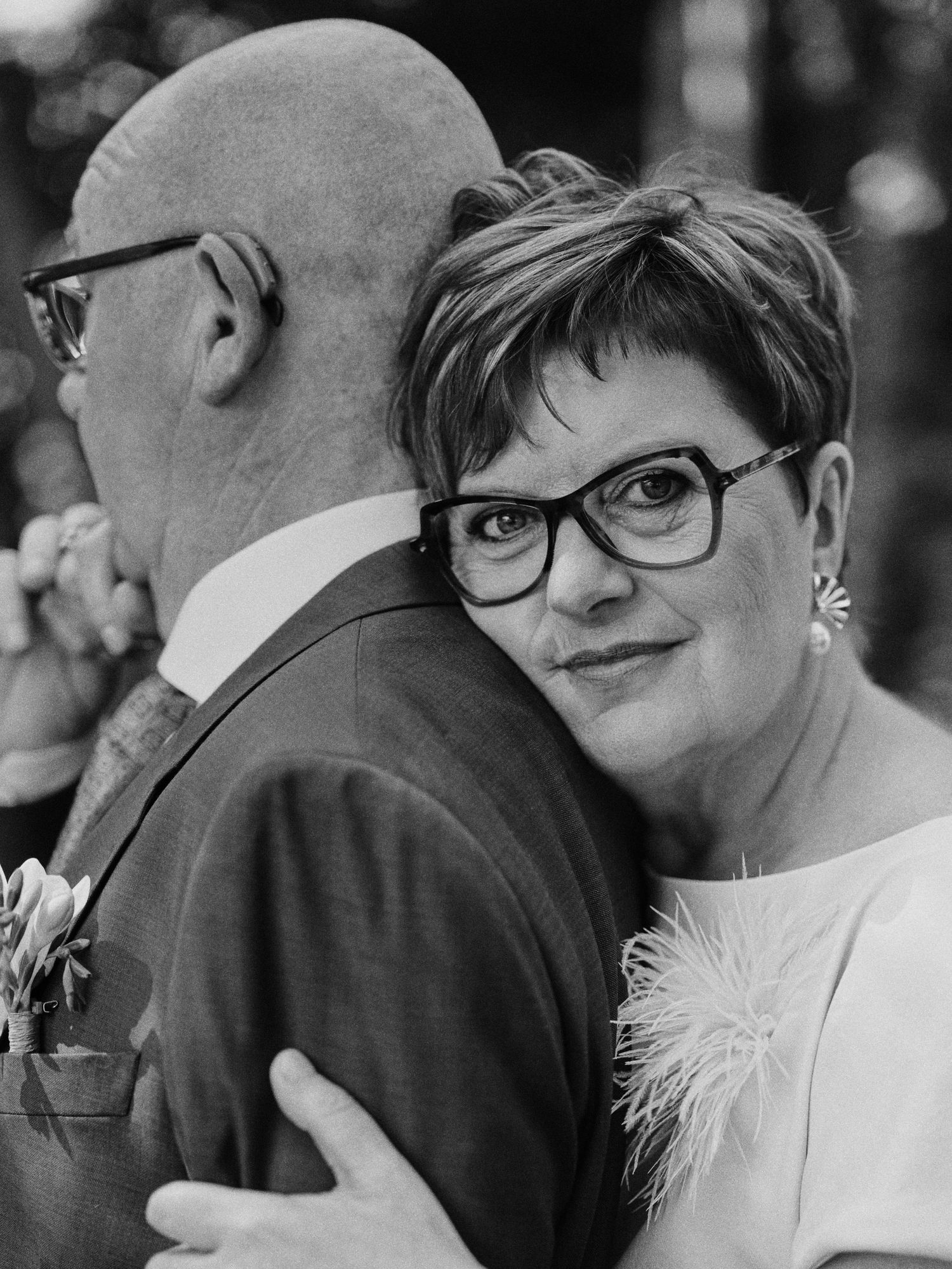 Black and white photo of a woman with short hair and glasses hugging a man in a suit at a wedding.