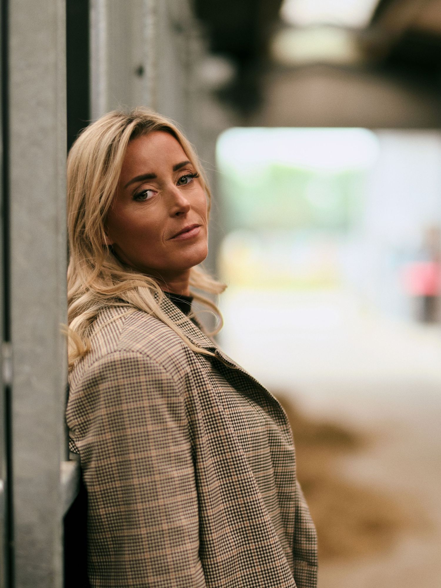 Blonde woman in houndstooth coat leaning against a wall, looking over her shoulder with a confident expression.