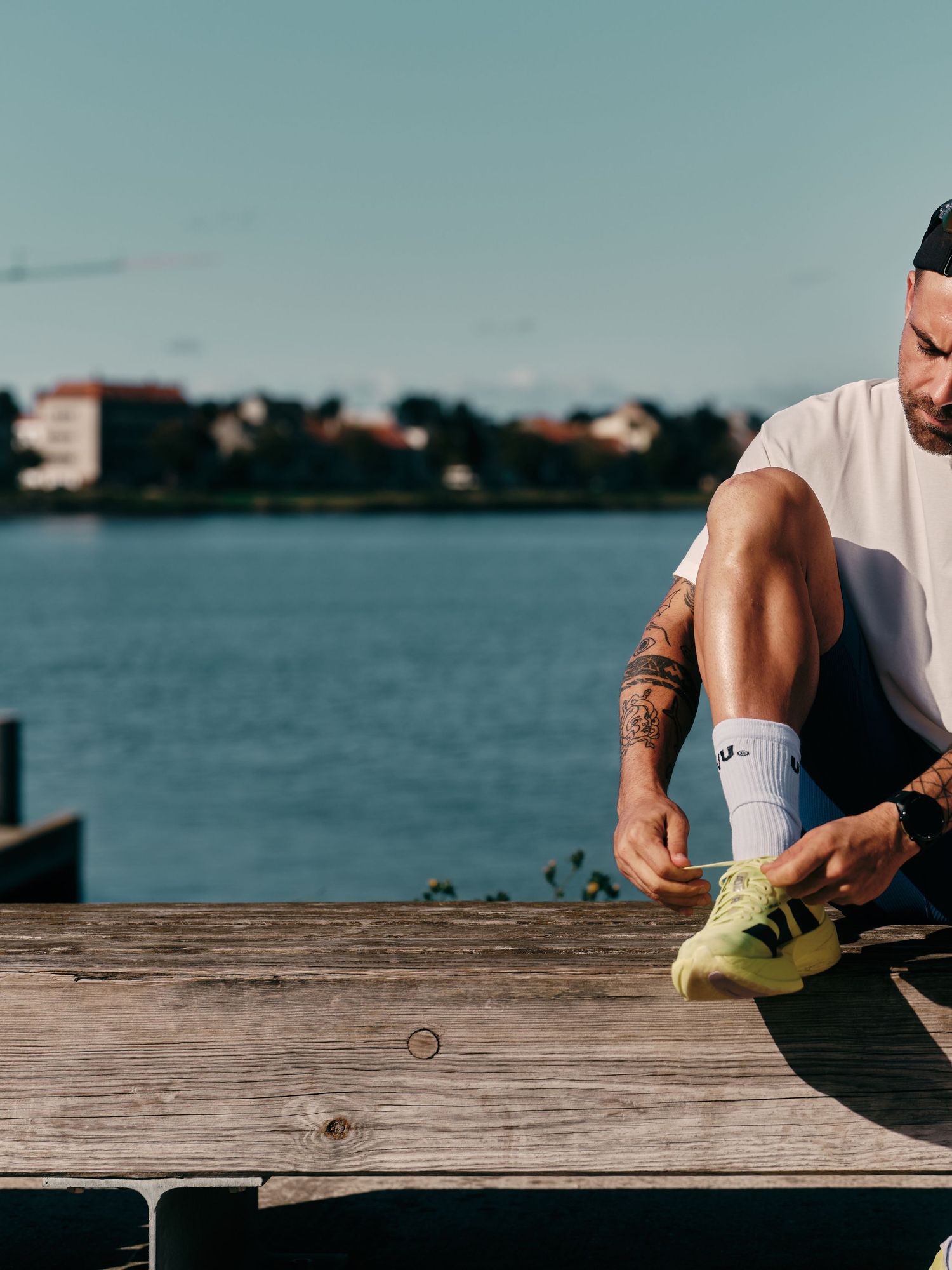 Tattooed man in white tee tying neon yellow sneakers on wooden dock by waterfront on sunny day.