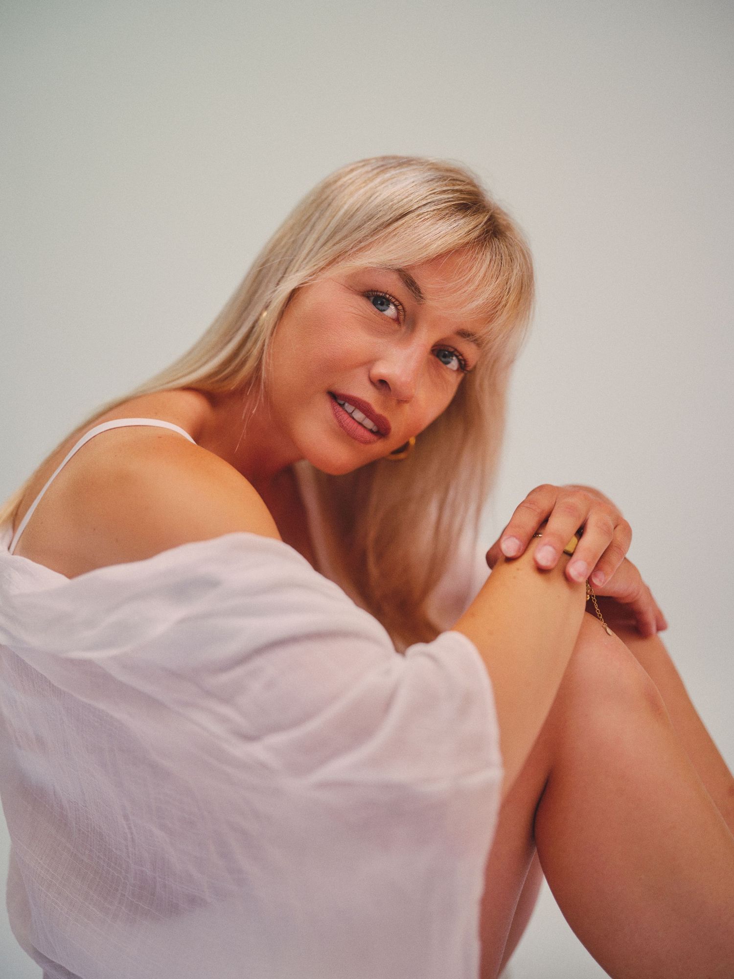 Blonde woman in white off-shoulder top sitting relaxed, looking at camera against light gray background.