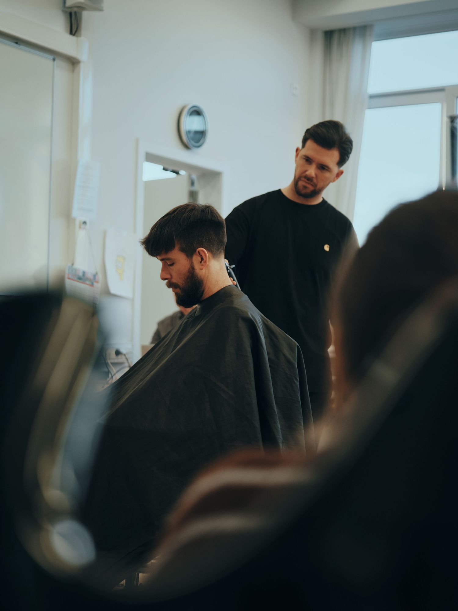 A barber in a black uniform cuts a bearded man's hair in a modern barbershop with bright windows.